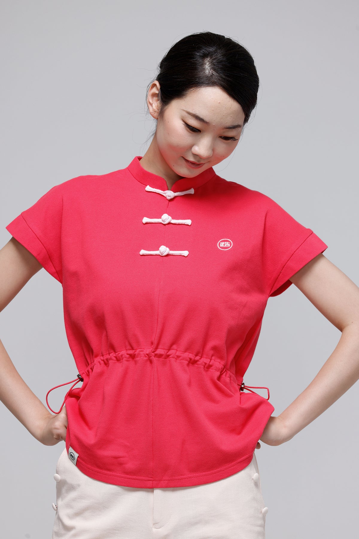 A woman wearing a bright pink short-sleeve top with three white Chinese-style frog closures and a drawstring waist, paired with off-white pants, against a solid gray studio background.
