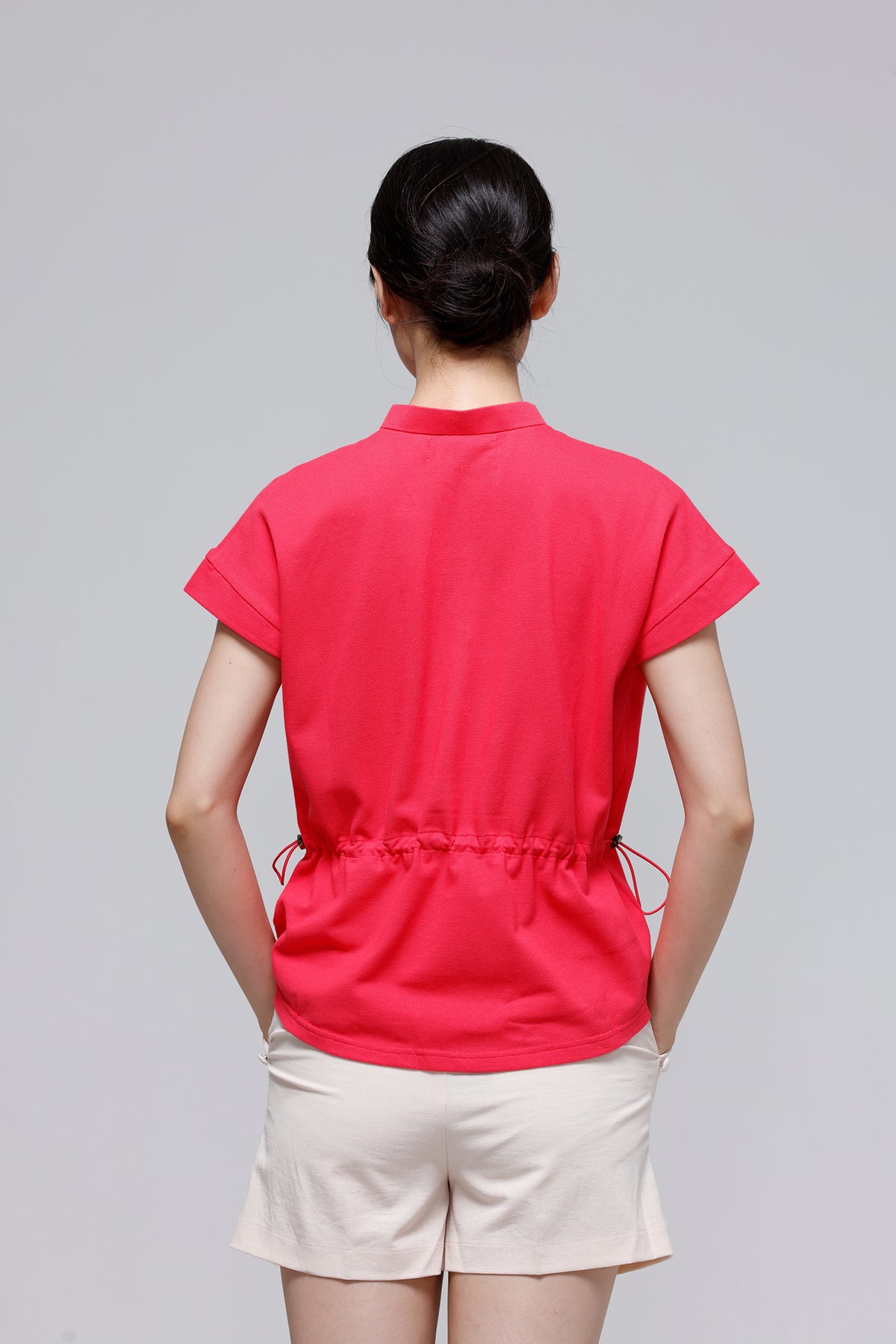 A woman viewed from behind wearing a bright pink short-sleeve top with a drawstring waist and light beige shorts, against a plain light gray studio background.