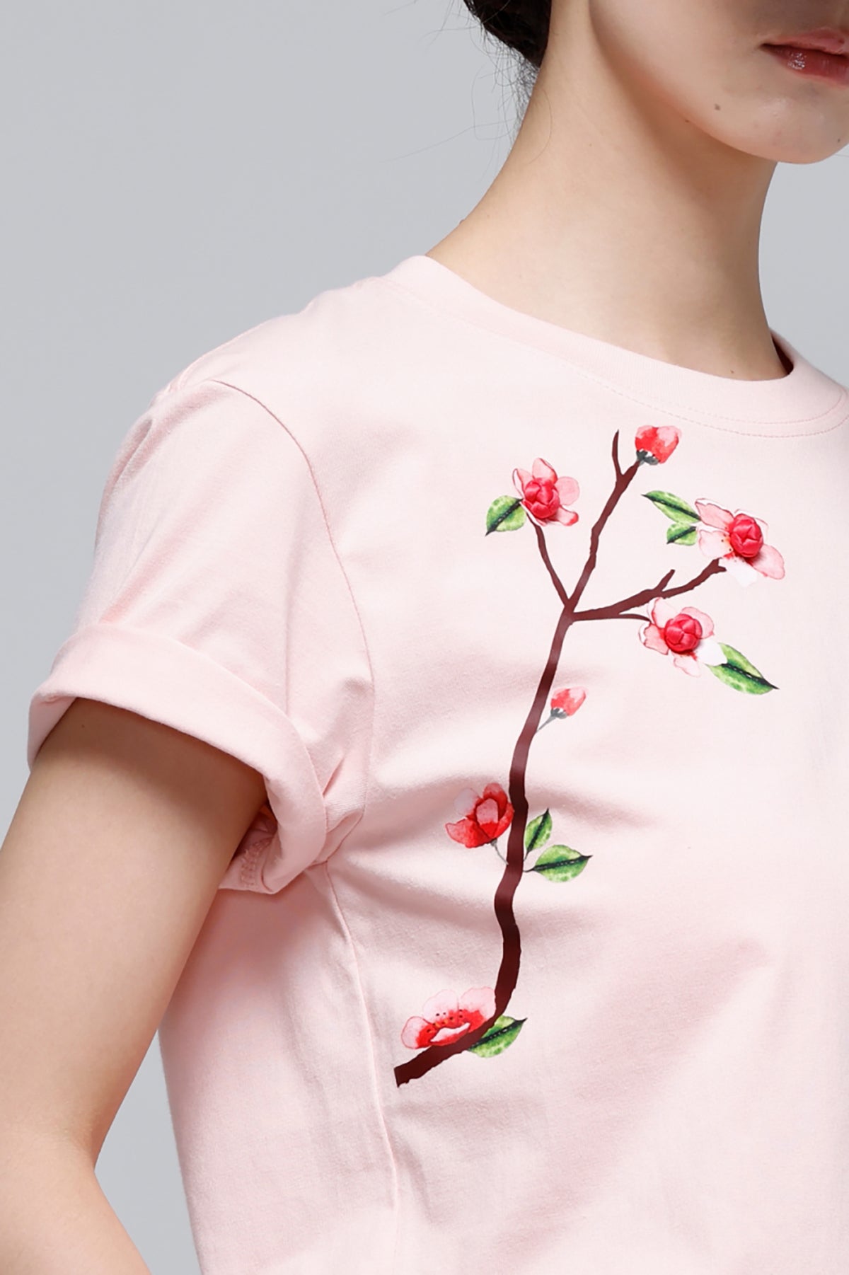 A close-up of a model wearing a light pink short-sleeve t-shirt with a watercolor-style embroidered cherry blossom branch graphic on the front.