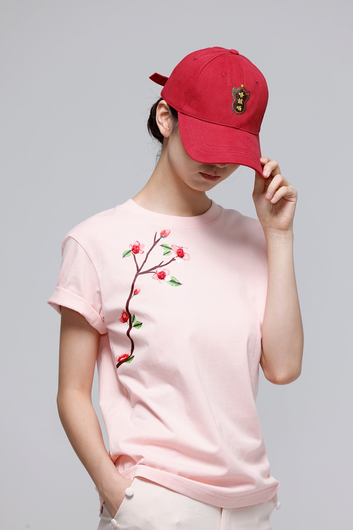 A model wearing a light pink crewneck t-shirt with a cherry branch embroidery on the front, paired with a red baseball cap and white pants, set against a solid gray studio background.