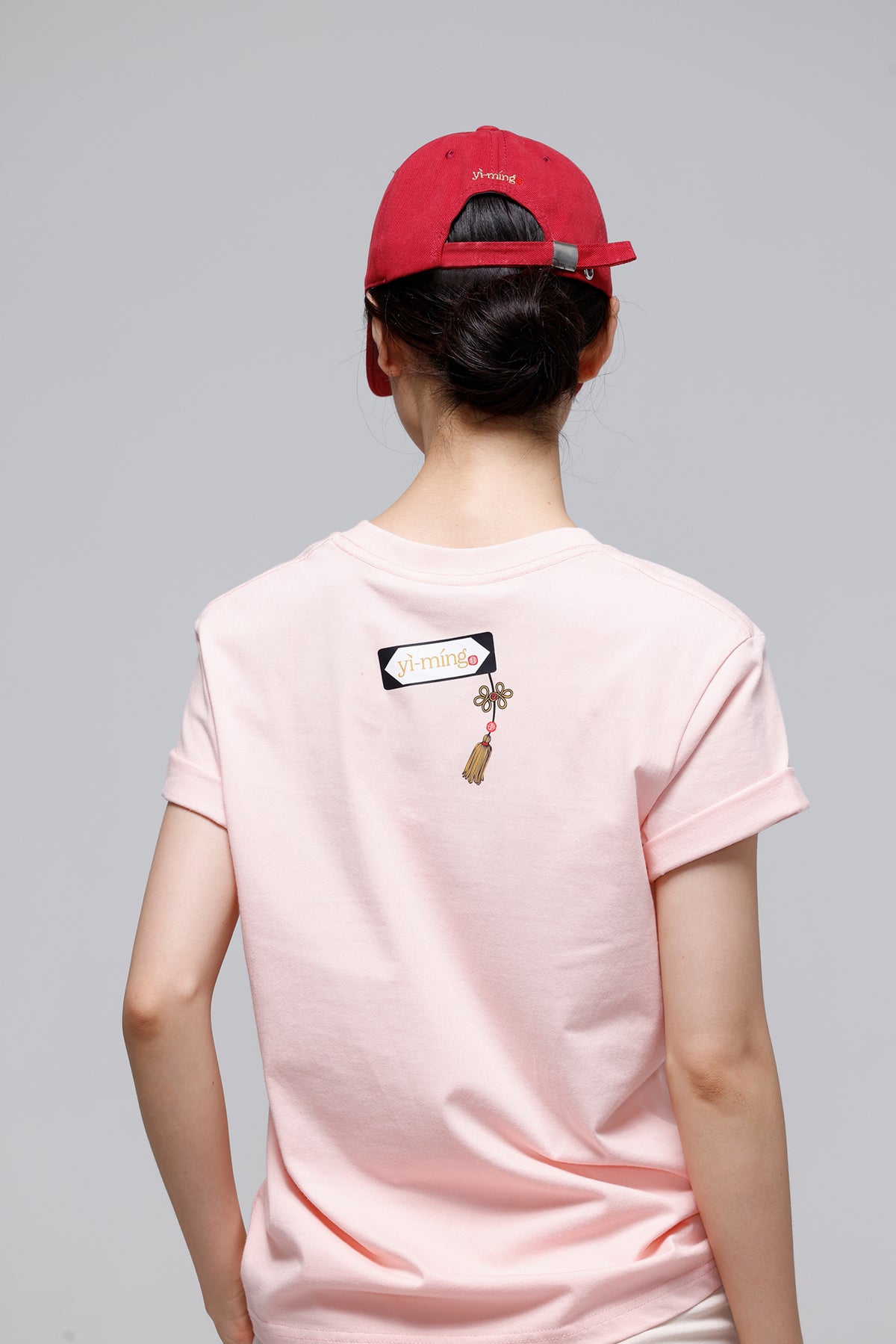A person shown from behind wearing a light pink t-shirt with a small graphic on the upper back and a red baseball cap, against a solid gray studio background.
