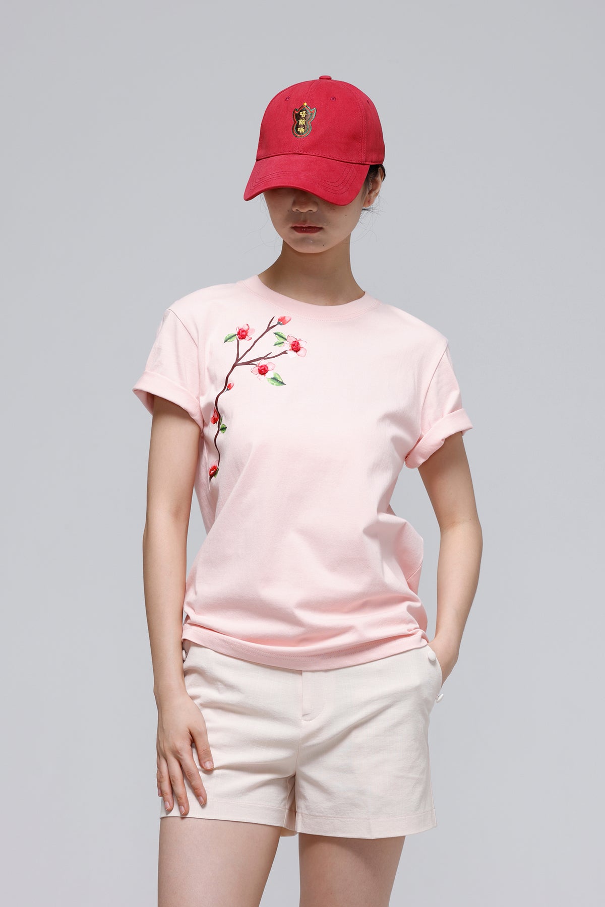 A model wearing a light pink short-sleeve t-shirt with a cherry branch embroidery on the chest, paired with off-white shorts and a red baseball cap, set against a solid gray studio background.