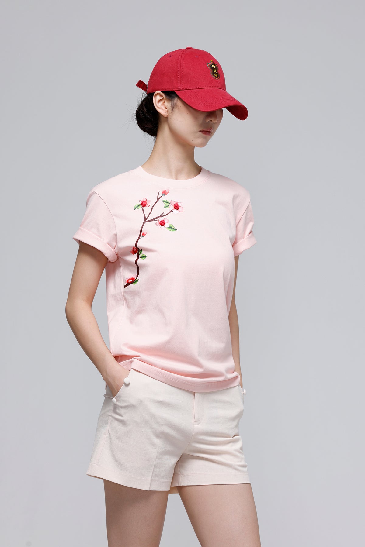 A woman modeling a light pink short-sleeve t-shirt with a cherry blossom branch embroidery on the chest, paired with off-white shorts and a red baseball cap, against a solid gray studio background.
