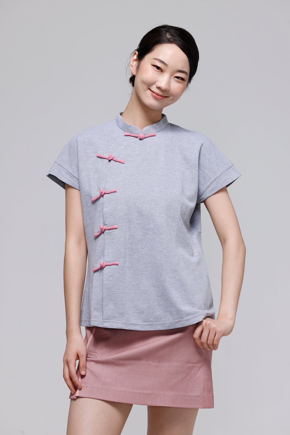 A woman modeling a light gray short-sleeve top with pink Chinese-style frog closures, paired with a dusty pink skirt, against a solid gray studio background.