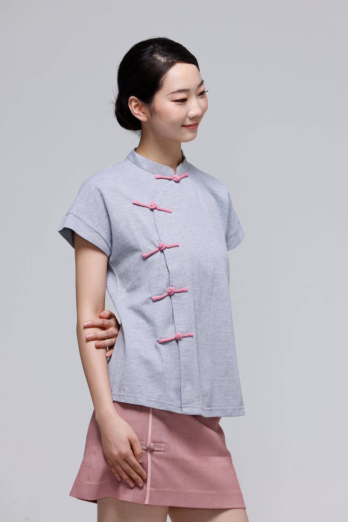 A woman modeling a light gray short-sleeve top with pink Chinese-style buttons, paired with a pink skirt, against a solid light gray background.