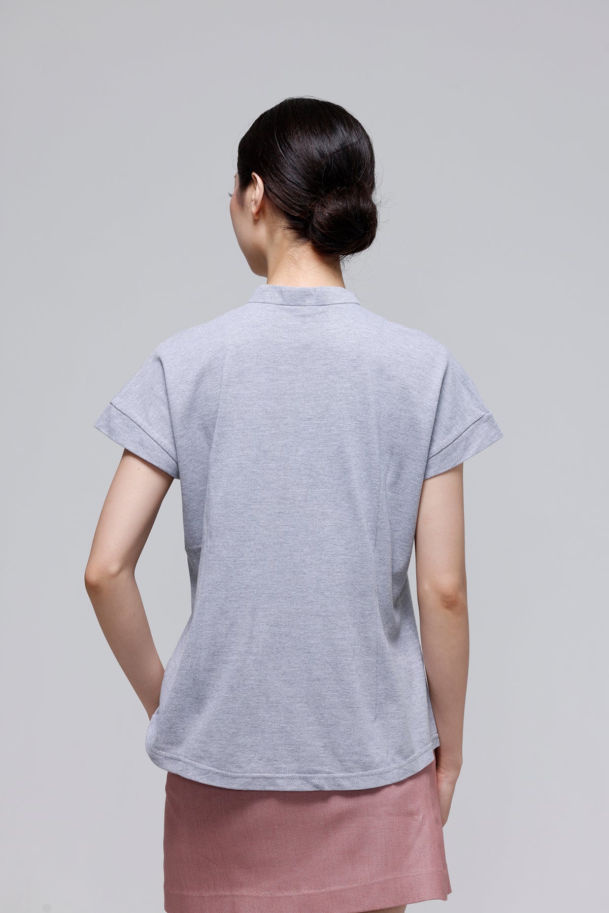 A woman viewed from behind wearing a light gray short-sleeve crewneck t-shirt and a dusty pink skirt, set against a solid light gray studio background.