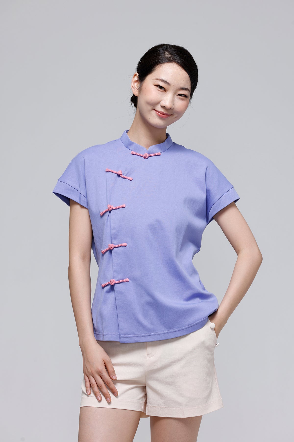 A woman modeling a light purple short-sleeve blouse with pink decorative buttons, paired with light beige shorts, against a solid light gray background.