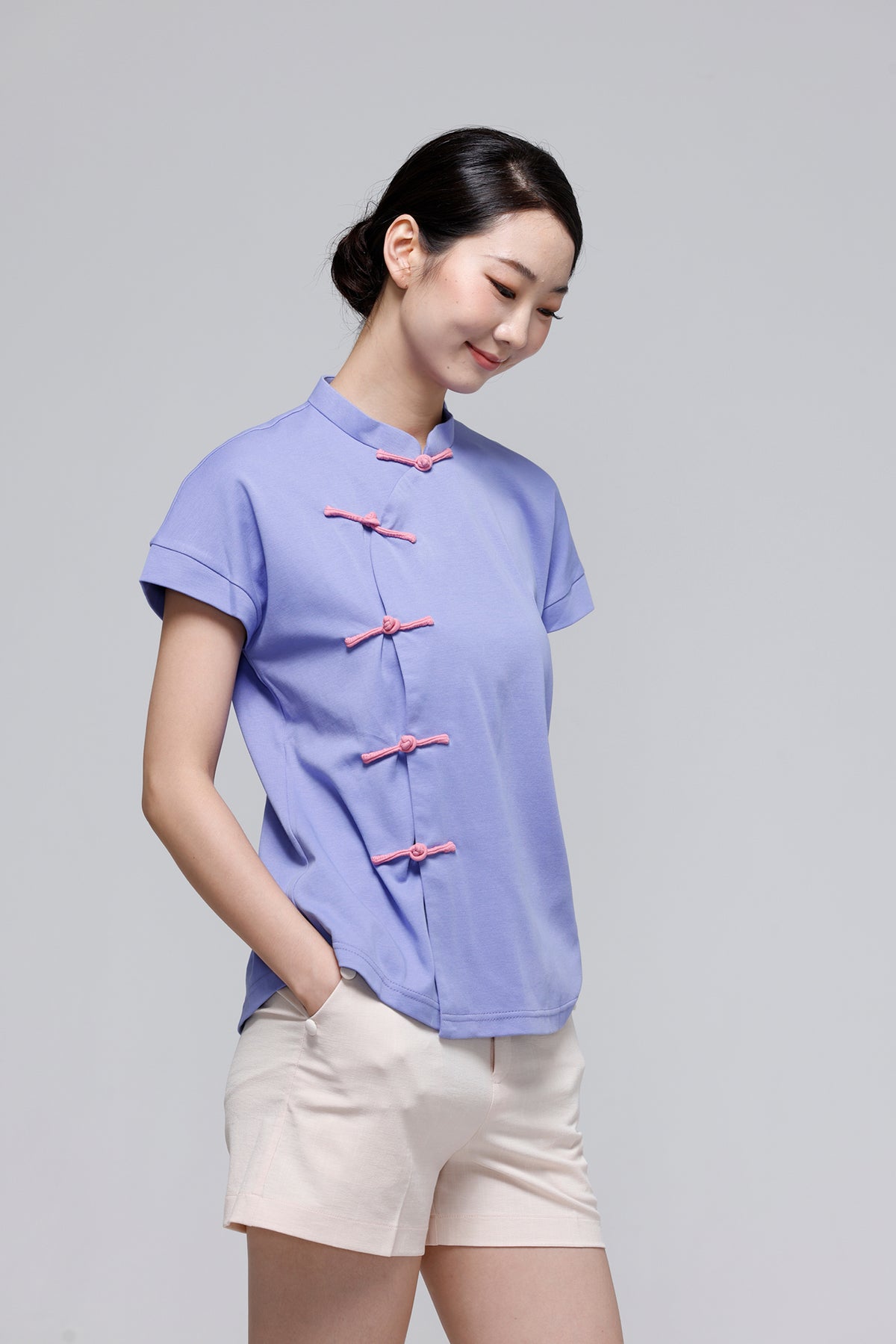 A woman wearing a light blue short-sleeve blouse with pink traditional Chinese frog closures and beige shorts, standing against a solid gray studio background.