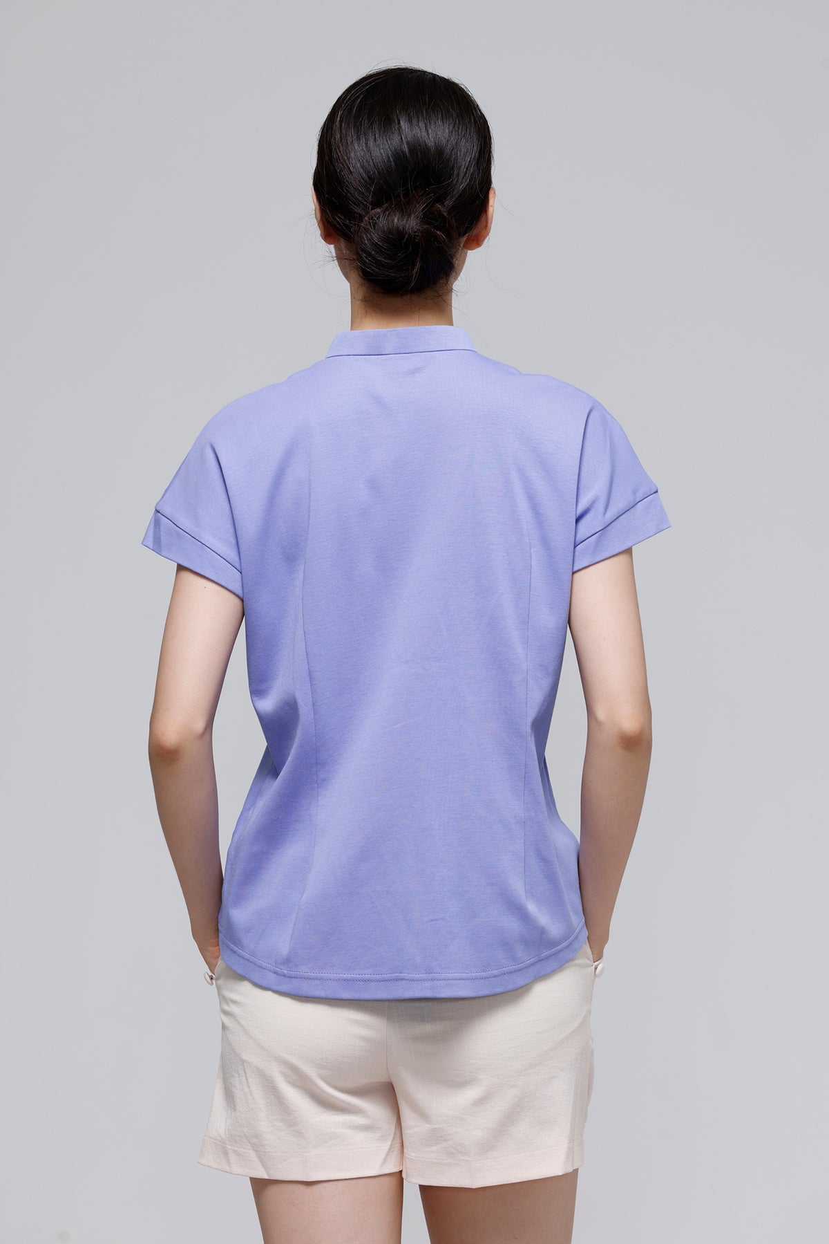A woman viewed from the back wearing a light lavender short-sleeve polo shirt and cream shorts against a plain light gray studio background.