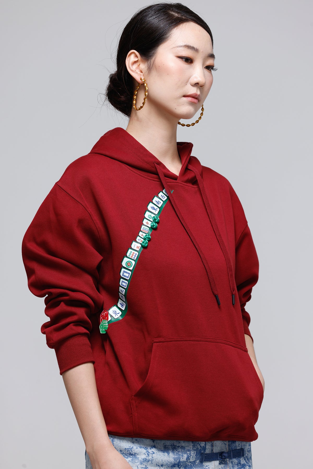 A woman wearing a deep red pullover hoodie with a green and white checkered patterned strap detail across the chest, paired with blue jeans, photographed against a solid gray studio background.