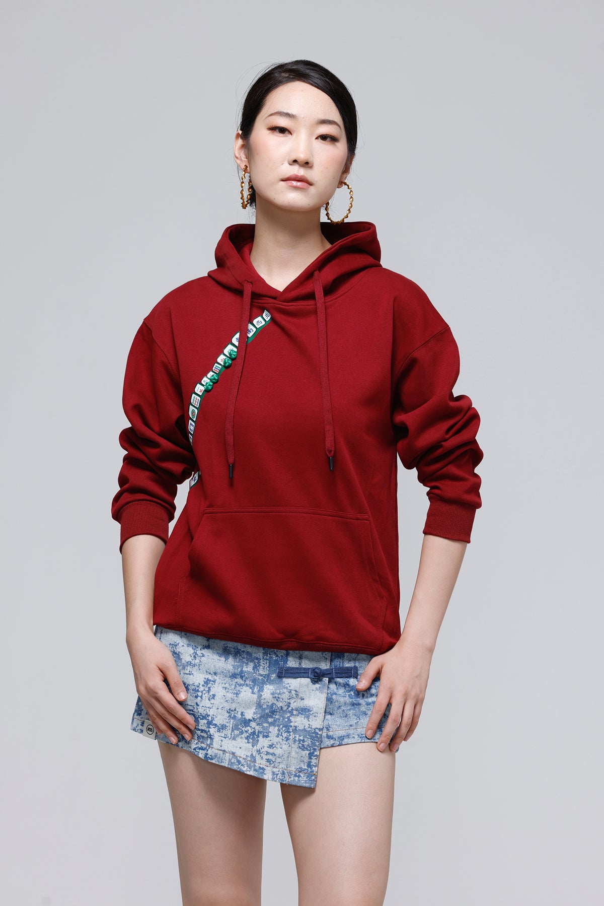 A woman modeling a dark red hoodie with a green and white graphic text detail on the left chest, paired with a blue patterned mini skirt, against a solid gray studio background.