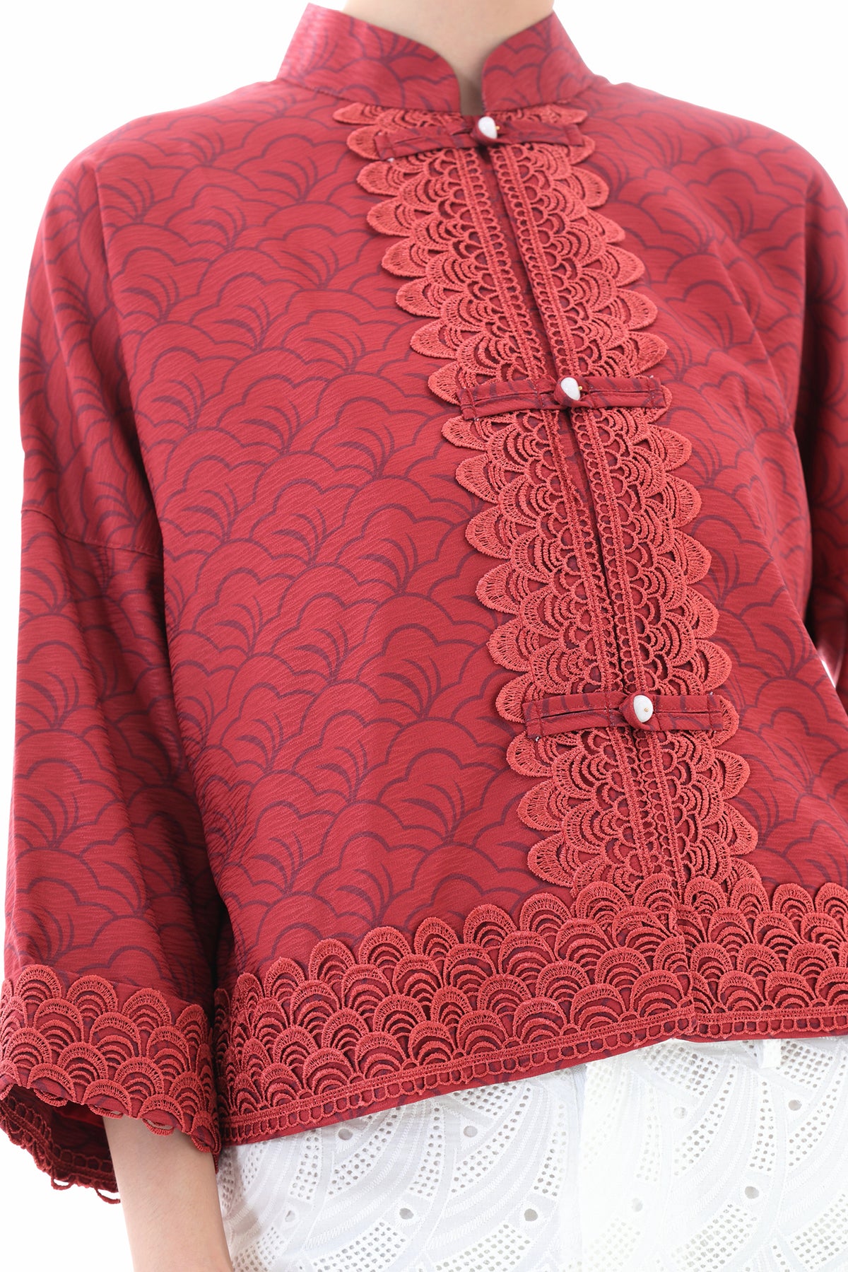 A close-up of a red cropped jacket with a scalloped lace trim down the front and matching scalloped hem, featuring three decorative buttons. The jacket is layered over a white lace top, shown on a model against a white background.