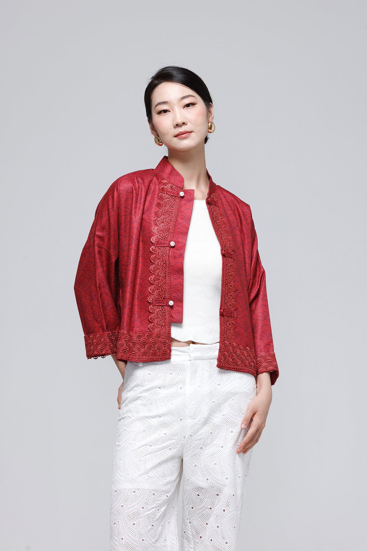 A woman modeling a red textured jacket with lace trim over a white top and white pants, photographed against a solid light gray studio background.