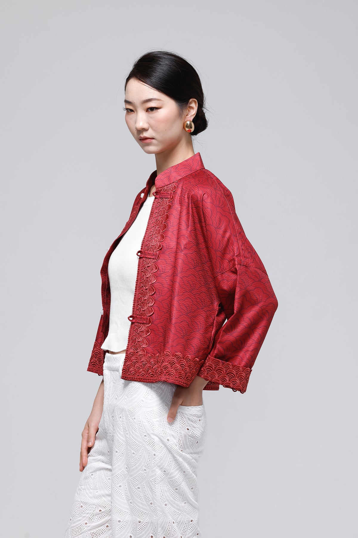 A woman modeling a red textured cropped jacket with traditional Chinese-style collar and button details, paired with a white top and white textured pants, against a solid light gray background.