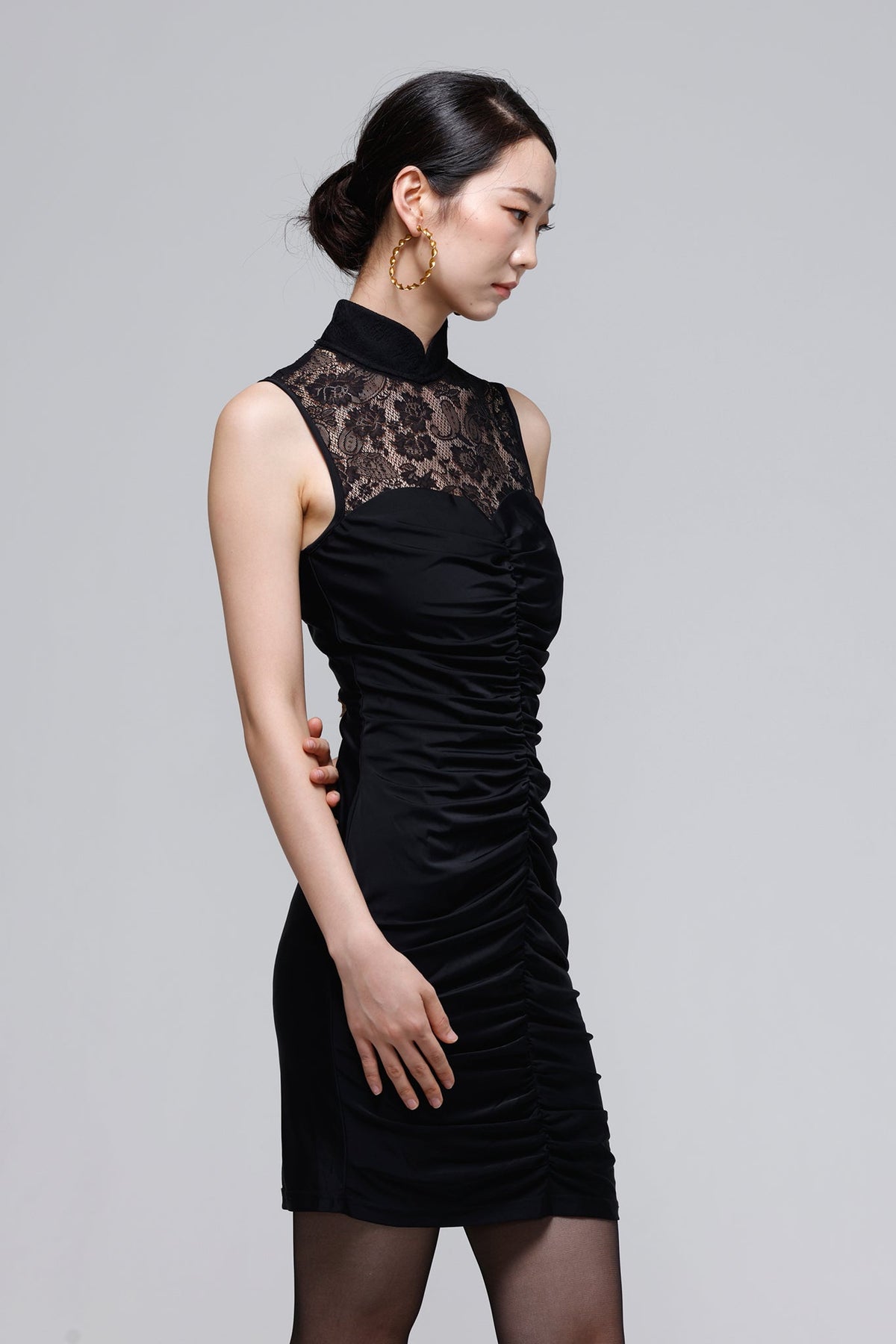 A woman modeling a sleeveless black dress with a high mock neck, black lace yoke, and ruched gathered skirt, photographed against a plain light gray studio background.