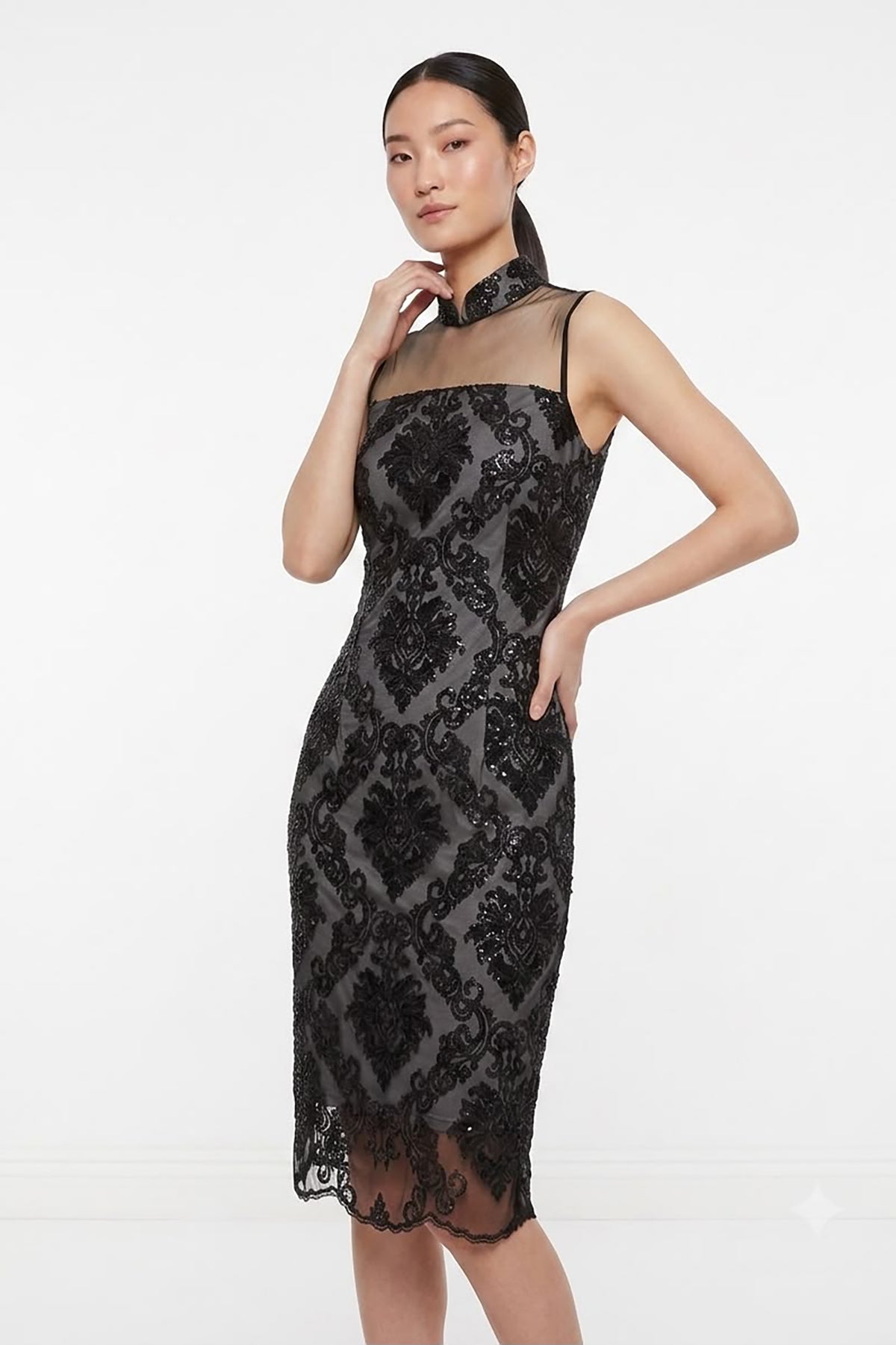 A woman modeling a sleeveless black lace midi dress with sheer mesh panels and a high neckline, photographed against a plain white studio background.