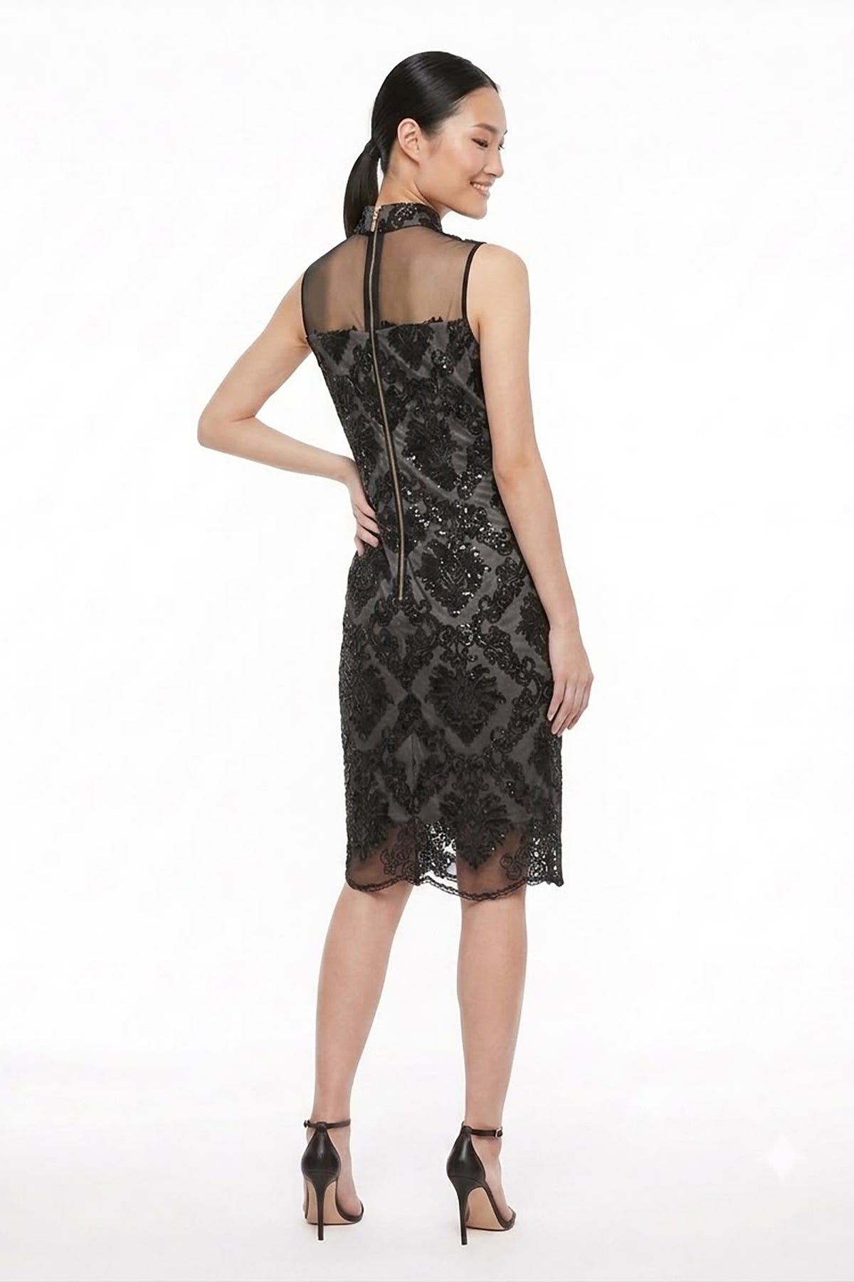A woman modeling a sleeveless black lace dress from the back, featuring a sheer mesh back panel and a high neckline, set against a plain white studio background.