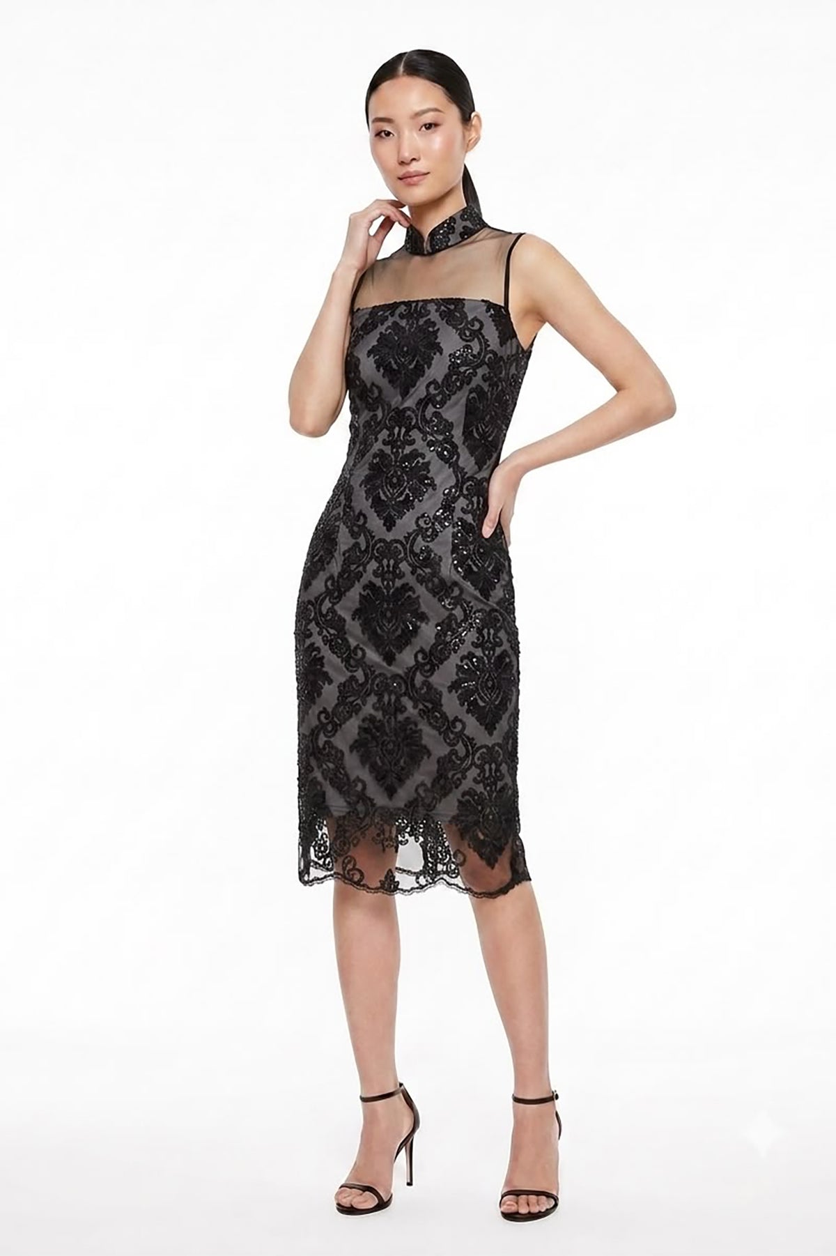 A woman modeling a black lace midi dress with a sheer high neck and thin shoulder straps, paired with black high-heeled sandals, against a plain white studio background.