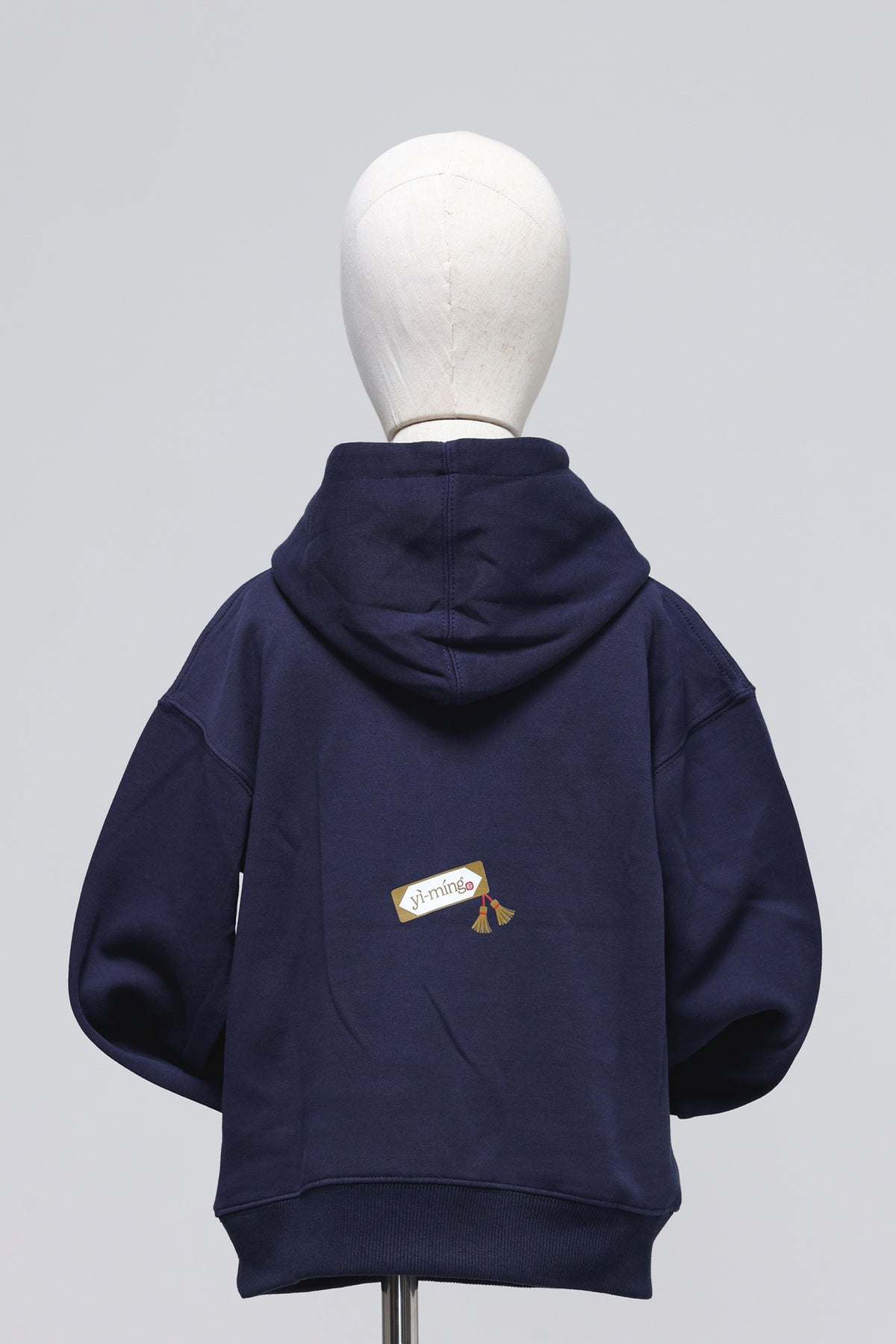 A navy blue hoodie shown from the back on a white mannequin head, featuring a small embroidered graphic on the lower back with the word 'amigo' and a tassel.