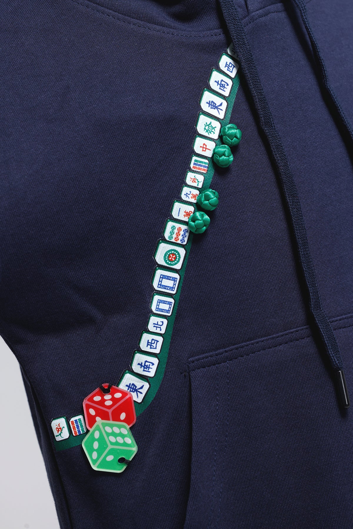 Close-up of a navy blue hoodie featuring a decorative green drawstring strap embellished with white and green Mahjong tile graphics and red and green dice charms.