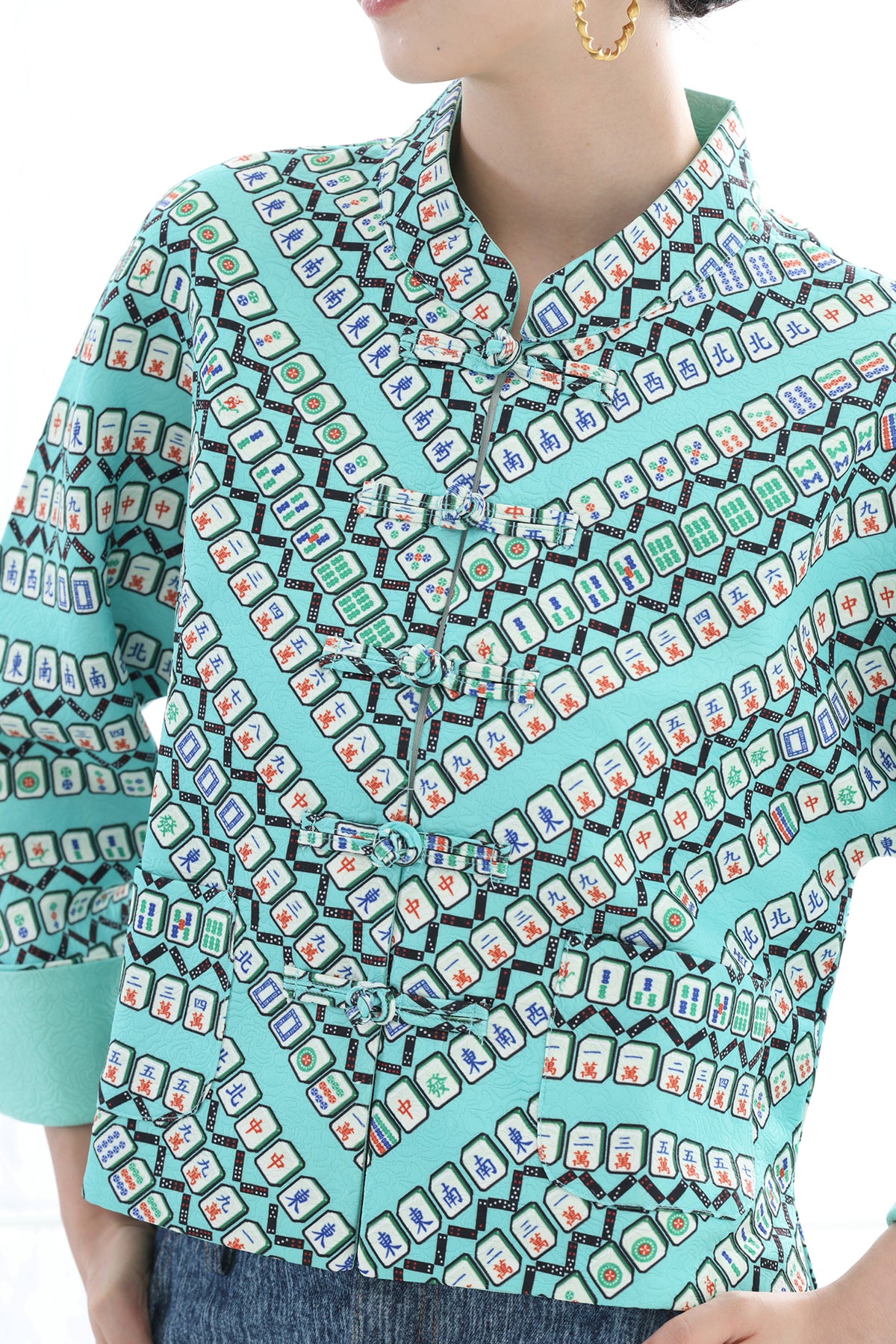 A close-up of a person wearing a turquoise collared button-up shirt with a dense geometric and card-like print in black, white, red, and blue. The image is cropped to show the torso, with a gold hoop earring visible on the left ear.