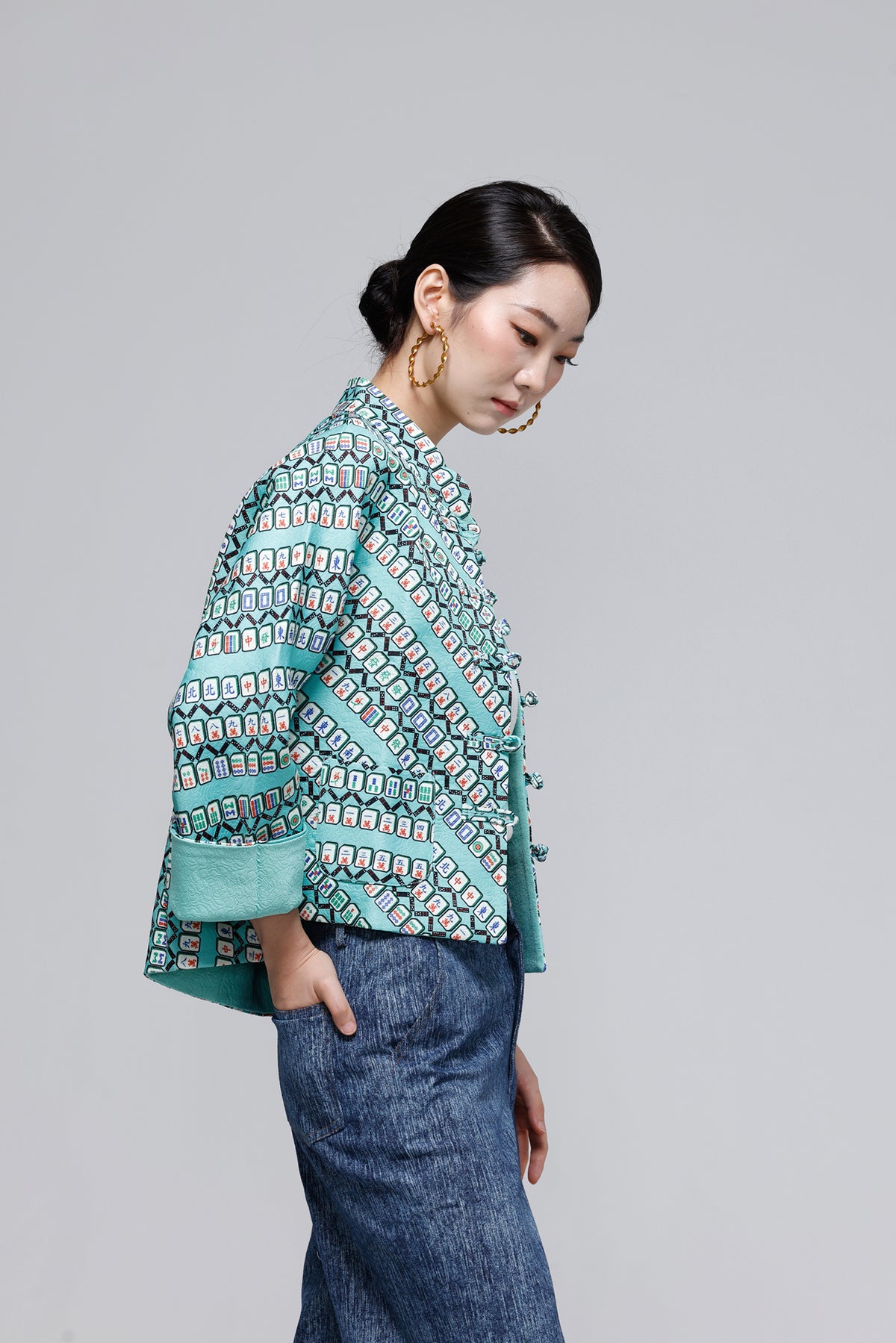 A woman wearing a mint green and blue patterned jacket with wide sleeves, paired with a long blue skirt, photographed against a solid gray studio background.