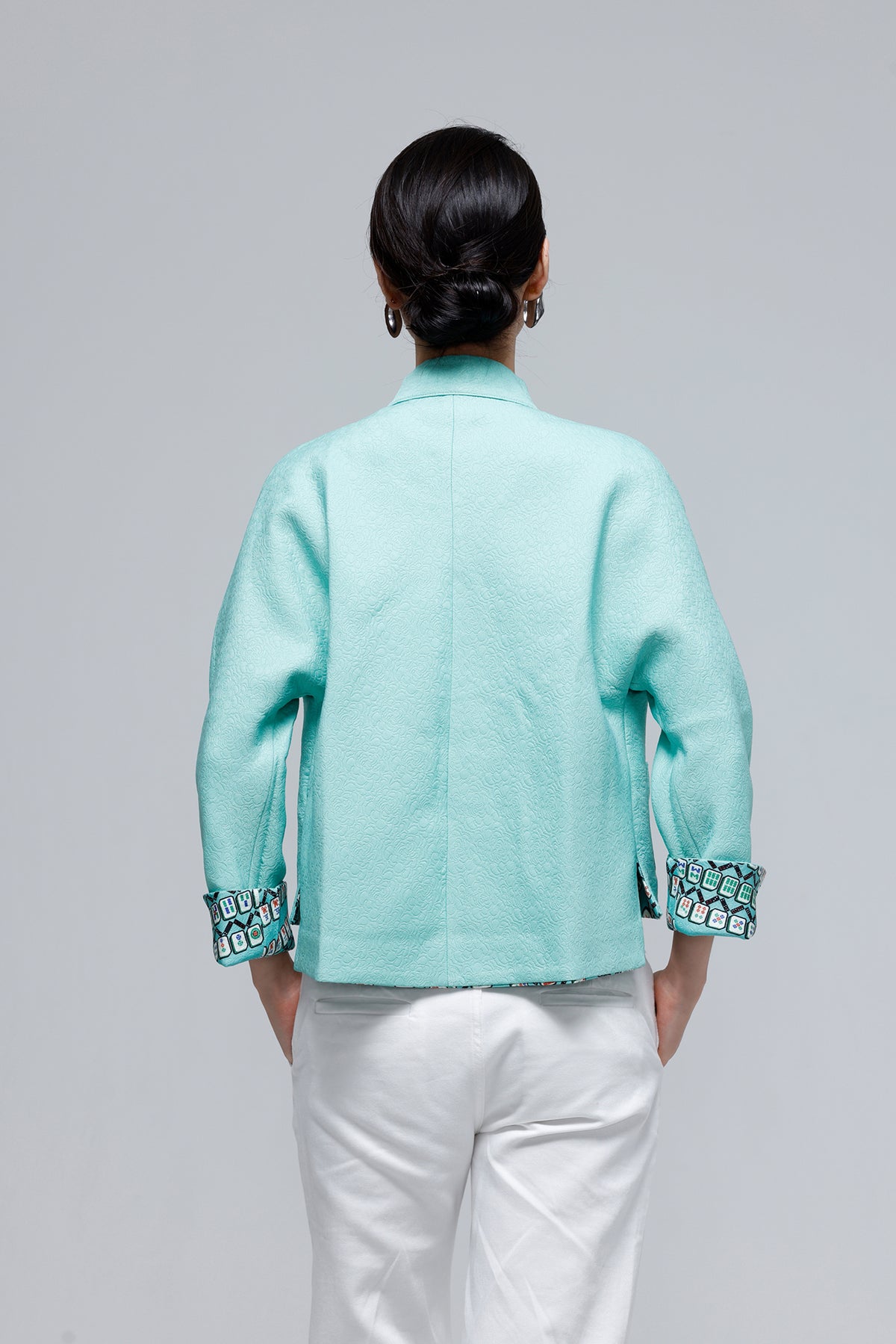 A woman shown from the back wearing a mint green cropped jacket with patterned cuff trim and white pants, photographed against a plain light gray studio background.