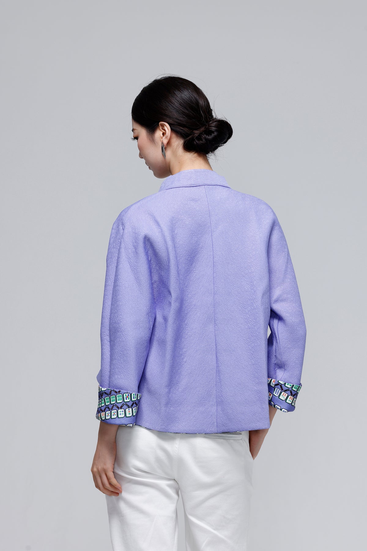A woman shown from the back wearing a lavender purple cropped blazer with patterned cuffs and white pants, photographed against a solid light gray studio background.