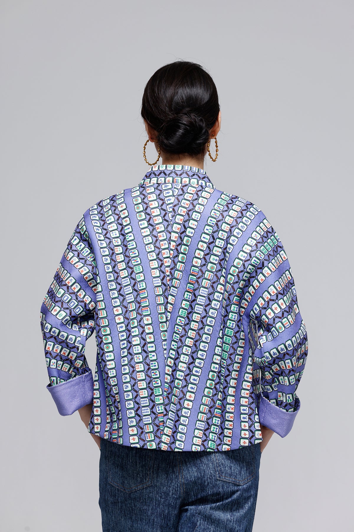 A woman viewed from behind wearing a purple patterned jacket with blue jeans, set against a solid gray studio background.