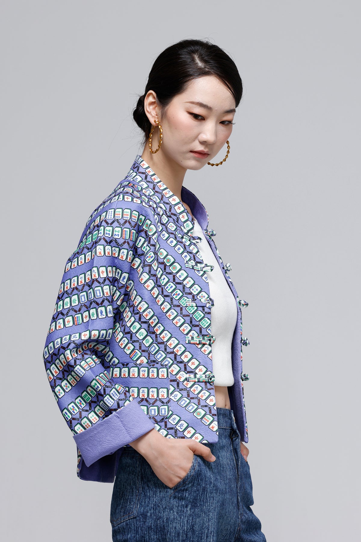 A woman wearing a purple cropped jacket with a colorful geometric pattern, styled with a white crop top and blue jeans against a solid gray background.