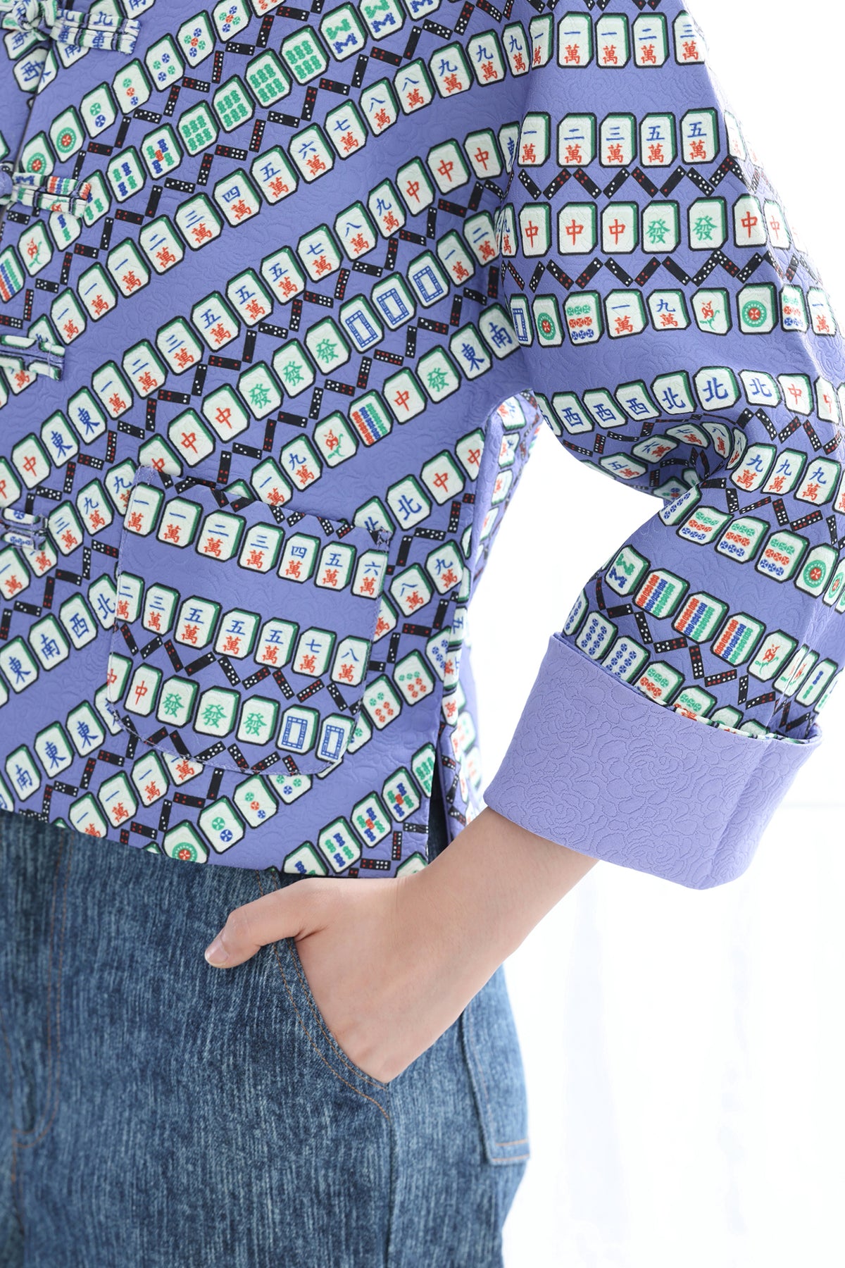 A close-up of a person wearing a lavender long-sleeve top with a repeating mahjong tile pattern in green, red, and white, paired with blue denim jeans.