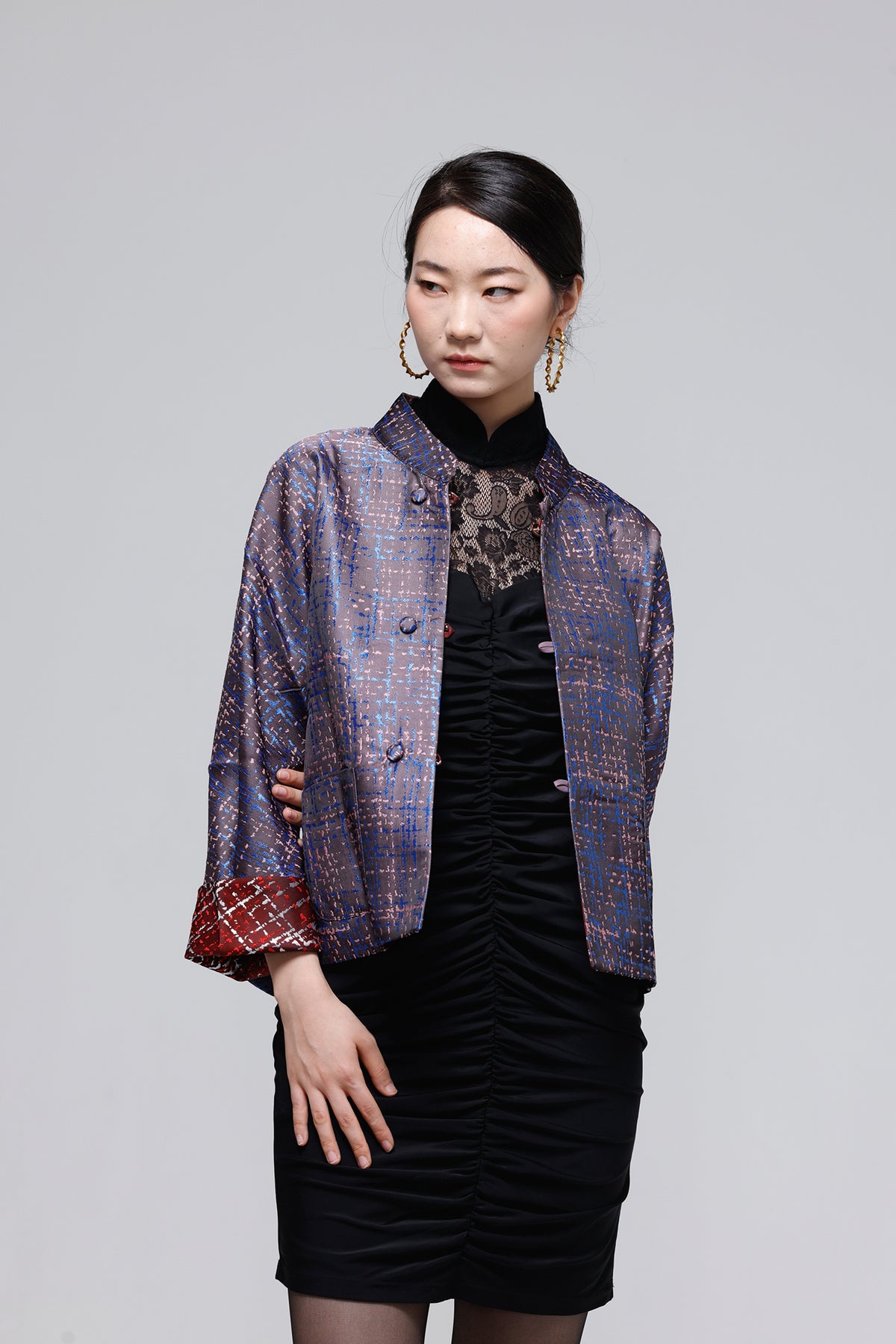 A woman modeling a short, cropped Chinese-style jacket with a blue and gold brocade pattern, worn over a black dress, against a solid gray studio background.
