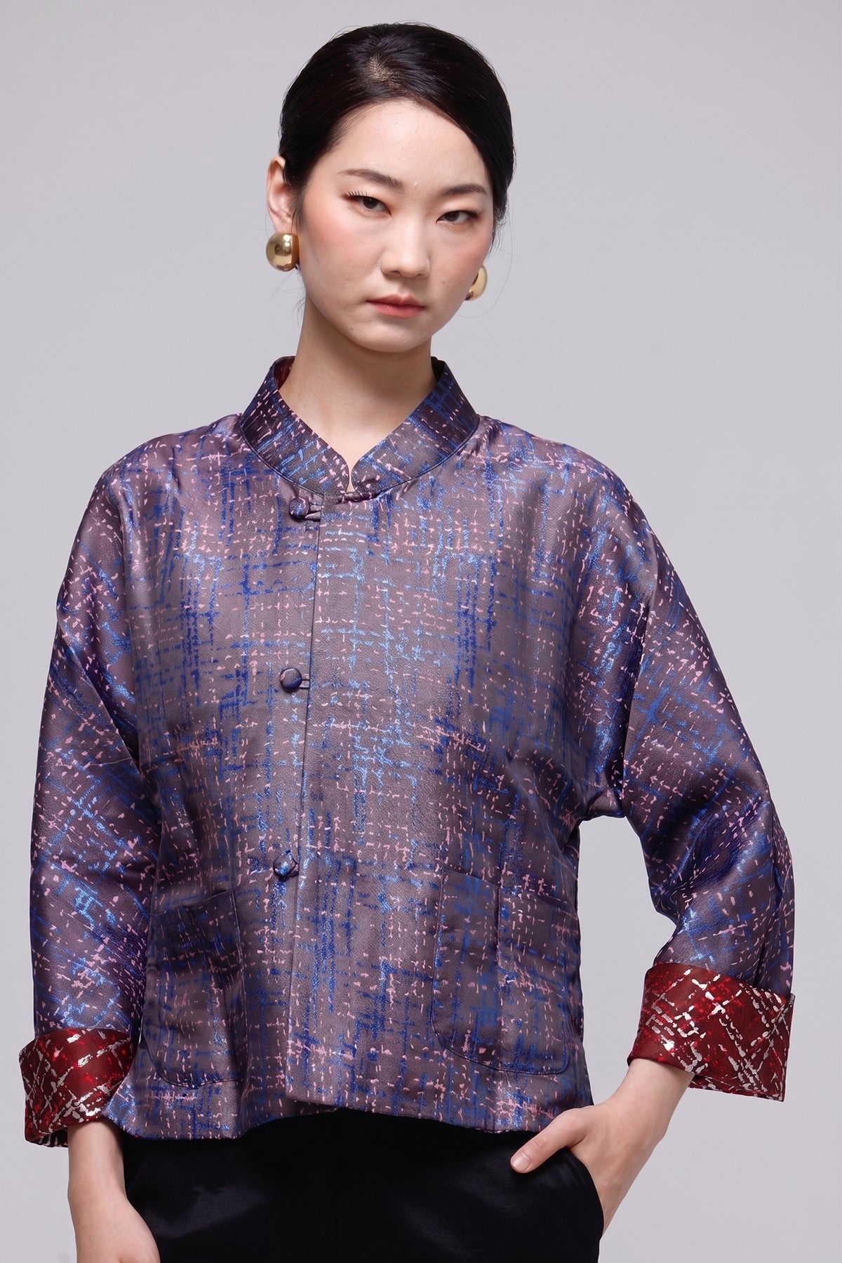 A woman modeling a short-sleeved, mandarin-collared button-up blouse with a textured, woven pattern in shades of blue, purple, and brown, featuring contrasting red patterned cuffs, set against a solid gray studio background.