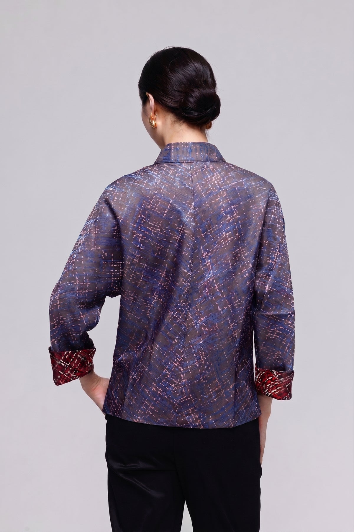 A woman viewed from the back wearing a long-sleeve collared blouse with an abstract woven pattern in purple, blue, and red, paired with black pants, against a light gray studio background.