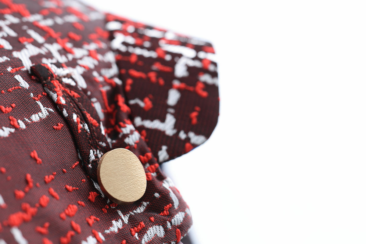 Close-up of a patterned fabric garment with a dark brown base, red and white abstract woven design, and a round gold-tone metal snap button.