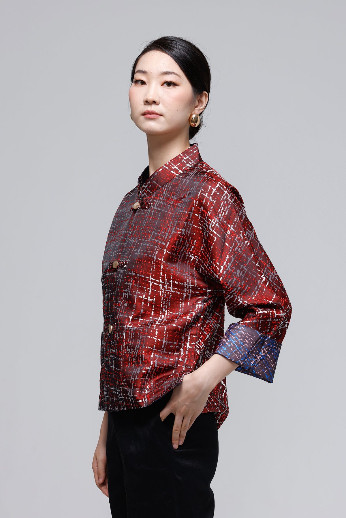 A woman modeling a red and black plaid button-up shirt with a subtle sheen, paired with black pants, against a solid gray studio background.
