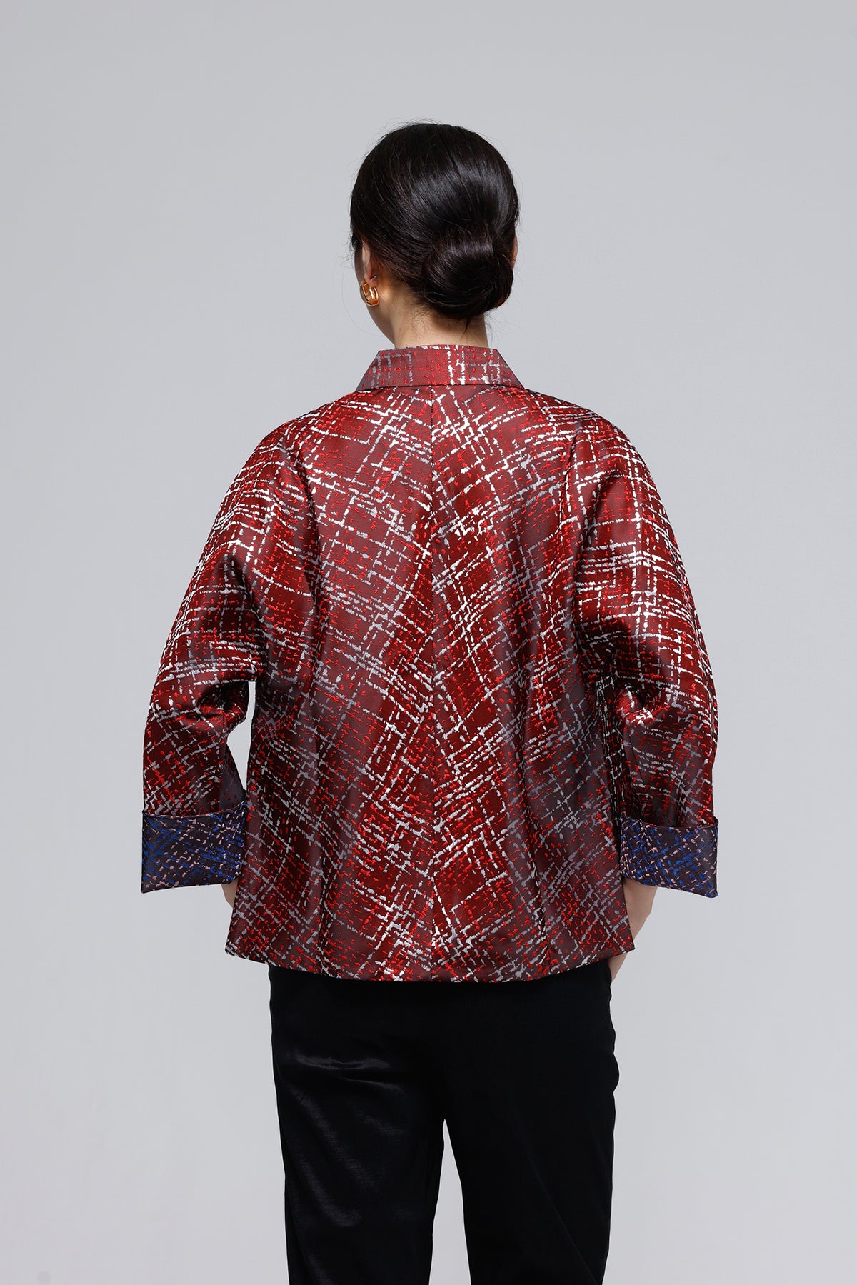 A woman viewed from behind wearing a red patterned long-sleeve shirt with black pants, set against a plain light gray studio background.