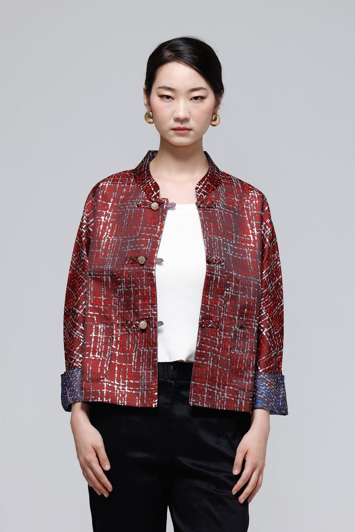 A woman modeling a red and black textured plaid jacket with a mandarin collar, button closure, and rolled cuffs, worn over a white top and black pants against a solid gray background.