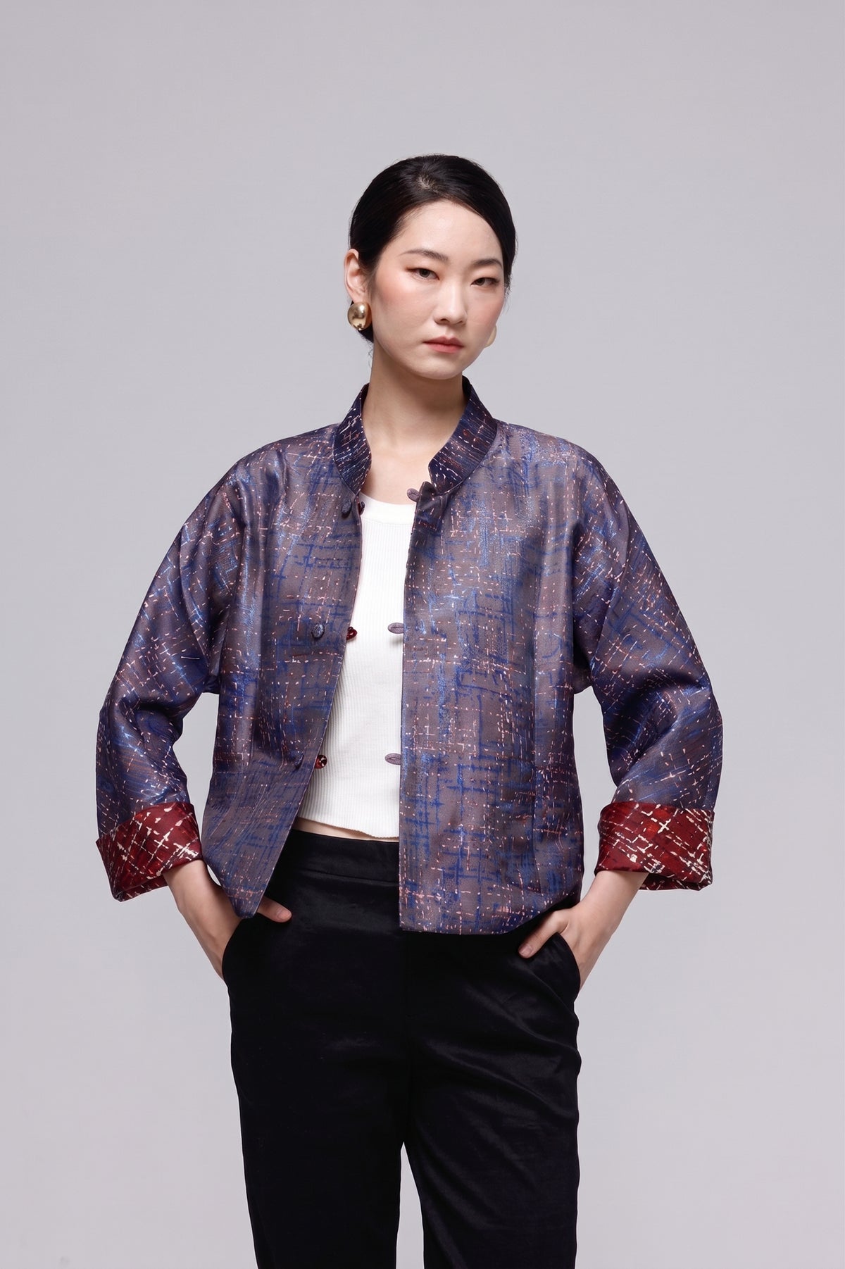 A model wearing a short-sleeved, mandarin-collared jacket with a blue and purple textured pattern, featuring red patterned cuffs and a white top underneath, paired with black pants, against a solid gray studio background.