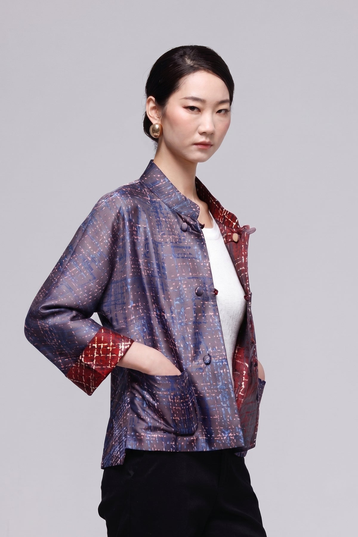 A woman modeling a short-sleeved, open-front jacket with a textured blue and purple patterned exterior and a red patterned lining, worn over a white top and black pants against a solid gray background.
