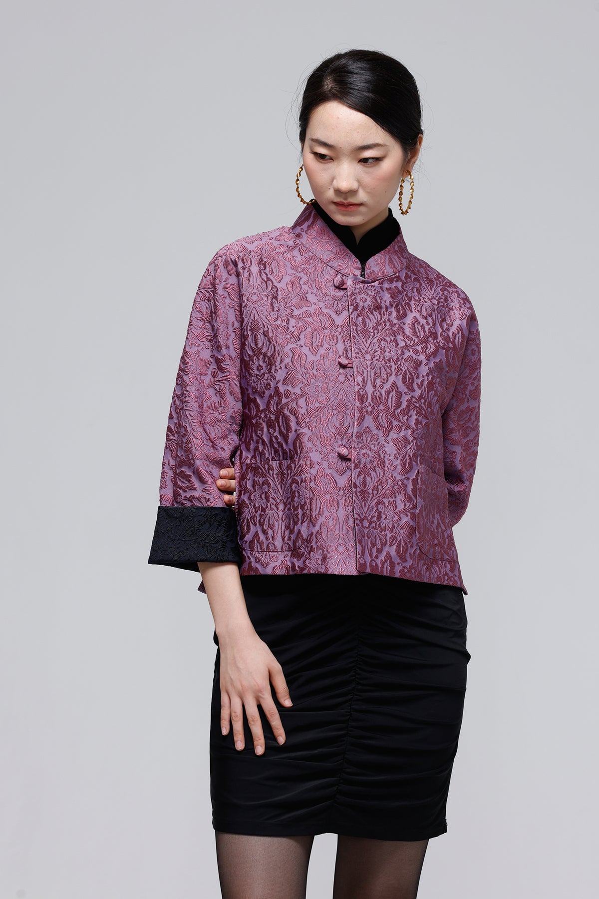 A woman modeling a short-sleeved, high-collar jacket in a textured mauve-purple fabric with black contrast cuffs, paired with a black ruched skirt, against a solid gray studio background.