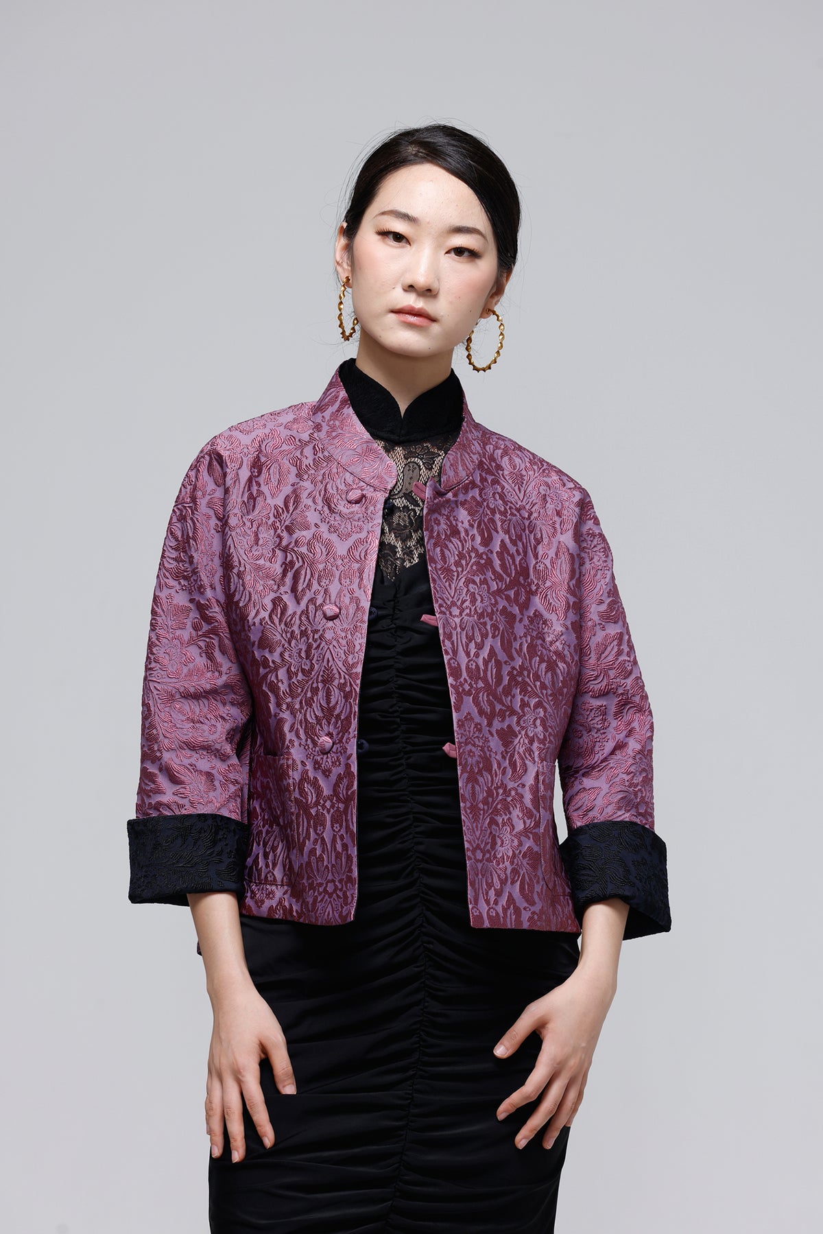 A woman modeling a short purple brocade jacket with black cuffs and collar, worn over a black dress, against a solid gray studio background.