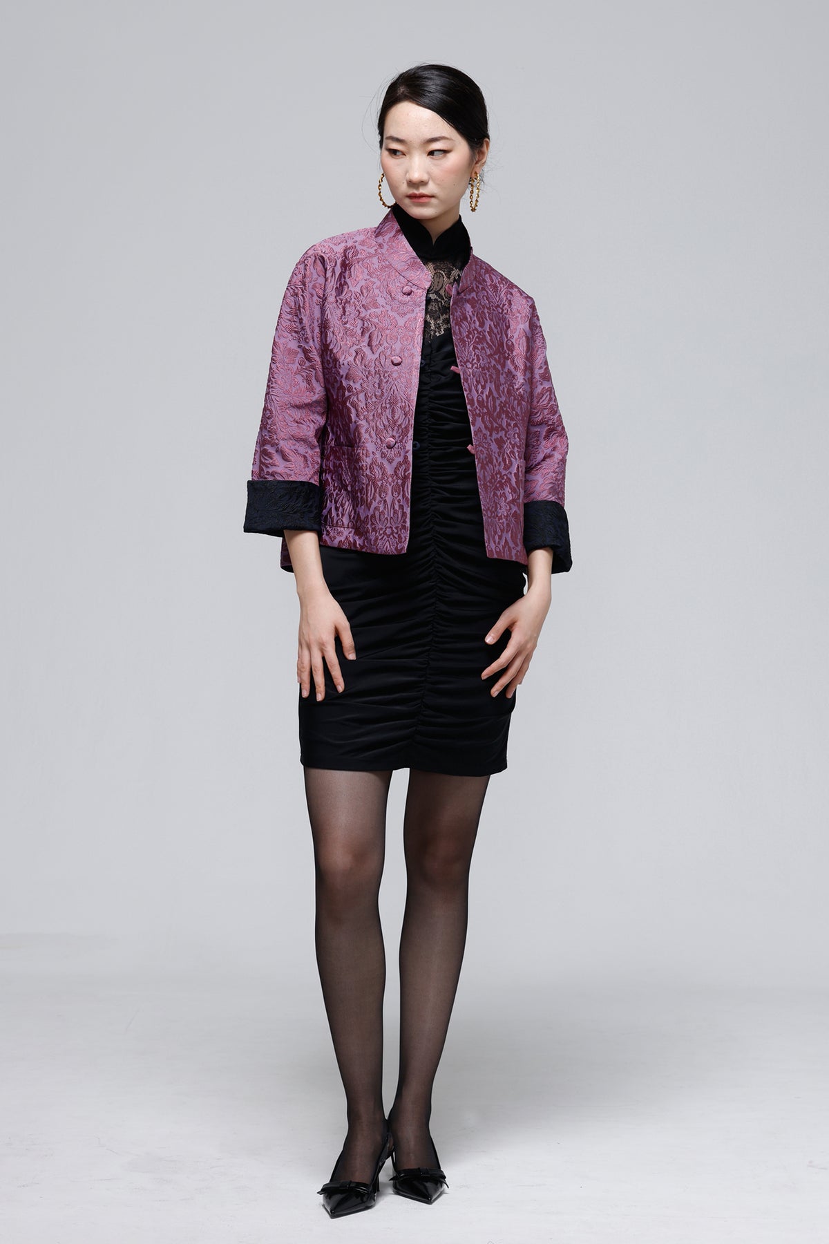 A model stands against a light gray studio background wearing a short purple patterned jacket with black cuffs over a black dress, black tights, and black heels.