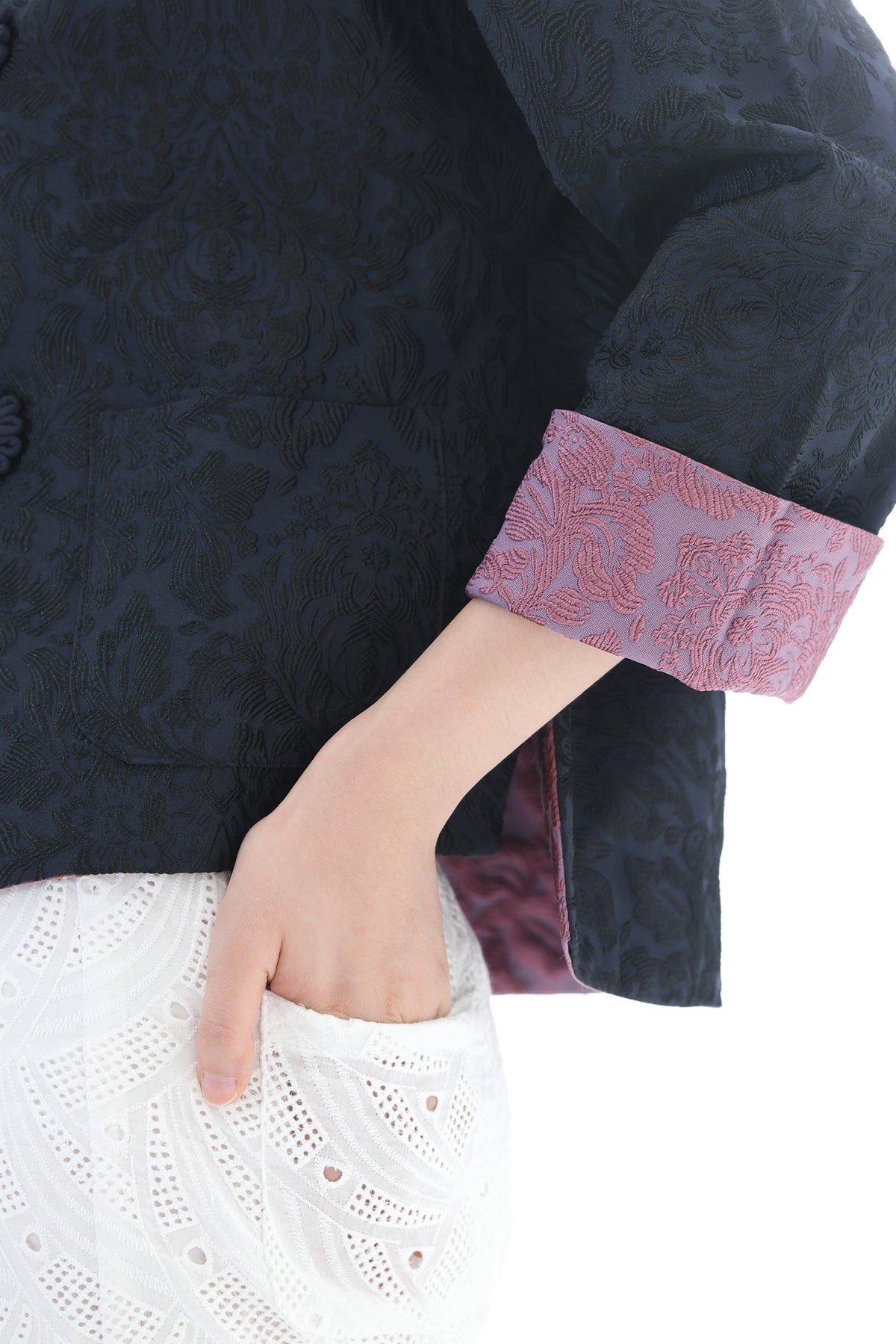 Close-up of a person wearing a dark navy textured jacket with a pink patterned cuff lining, paired with a white eyelet skirt, against a white background.