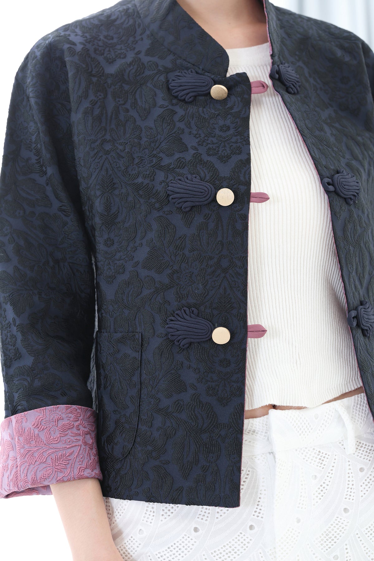 A close-up of a model wearing a dark navy blue brocade jacket with tassel-style buttons, layered over a white ribbed top and white skirt.