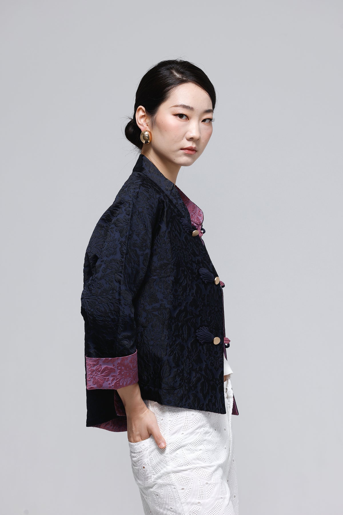A woman modeling a dark navy blue textured jacket with purple trim on the collar, cuffs, and front buttons, paired with white pants, against a solid gray studio background.