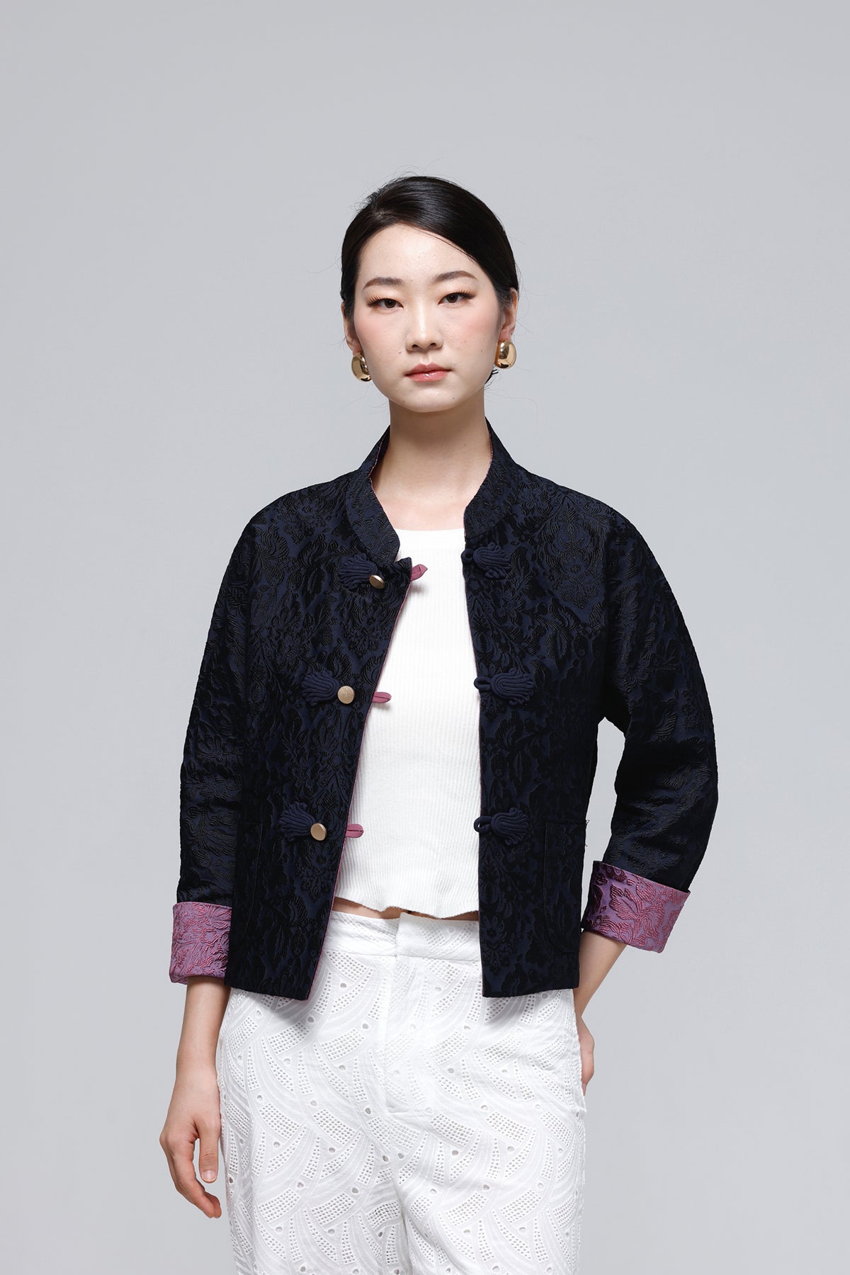 A woman modeling a dark navy textured jacket with purple cuff lining and gold buttons, worn over a white top and white pants, against a solid gray background.