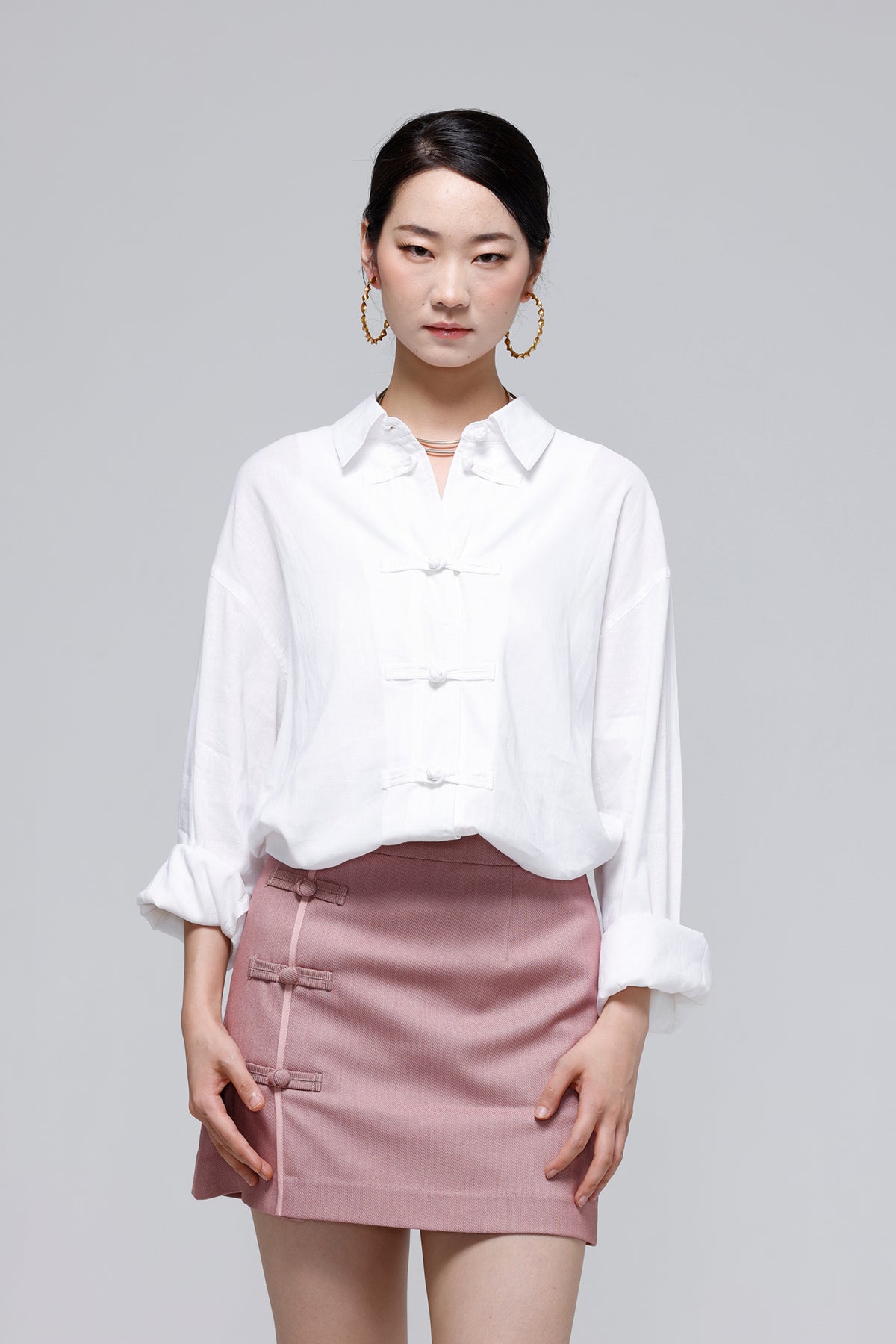 A woman modeling a white long-sleeve blouse with traditional Chinese frog closures and a dusty pink mini skirt with matching frog closures, set against a solid gray studio background.