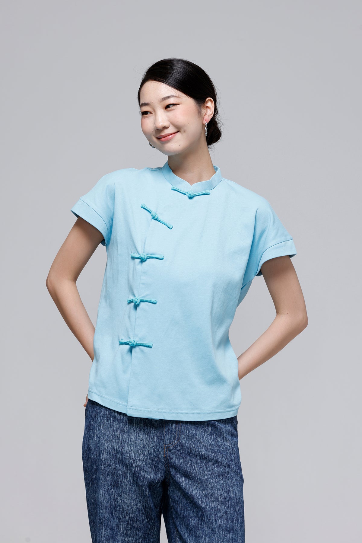 A woman modeling a light blue short-sleeve top with traditional Chinese-style frog closures, paired with dark blue textured pants, against a solid gray studio background.