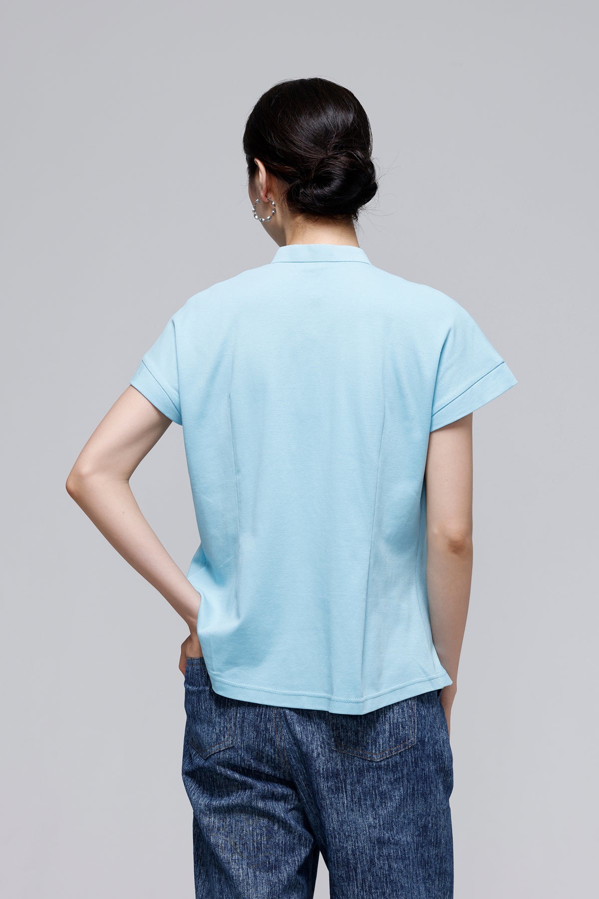A woman shown from the back wearing a light blue short-sleeve polo shirt and dark blue jeans against a solid gray studio background.